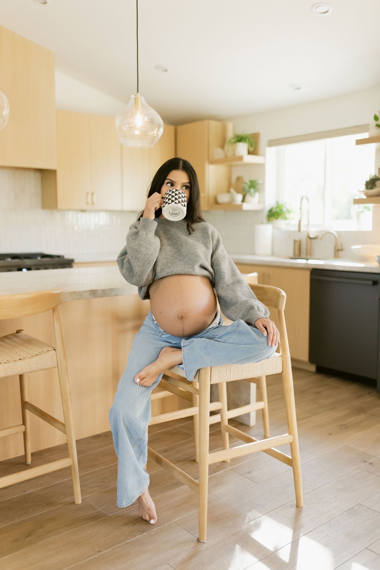 Pregnant woman sitting on a wooden bar stool in a modern kitchen, sipping from a mug with her baby bump exposed.