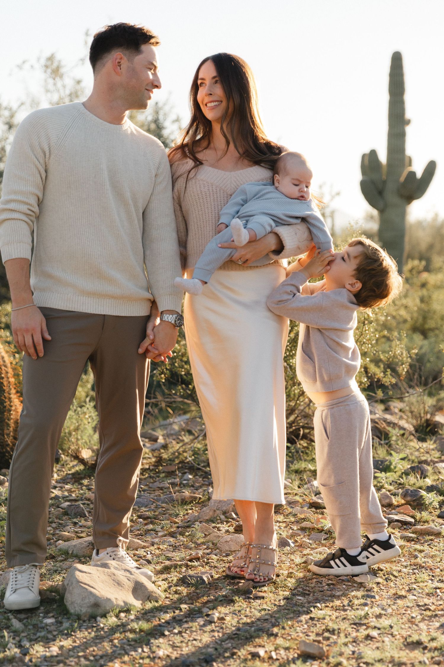 Family wearing coordinated neutral outfits stands together in desert landscape with saguaro cactus at sunset.