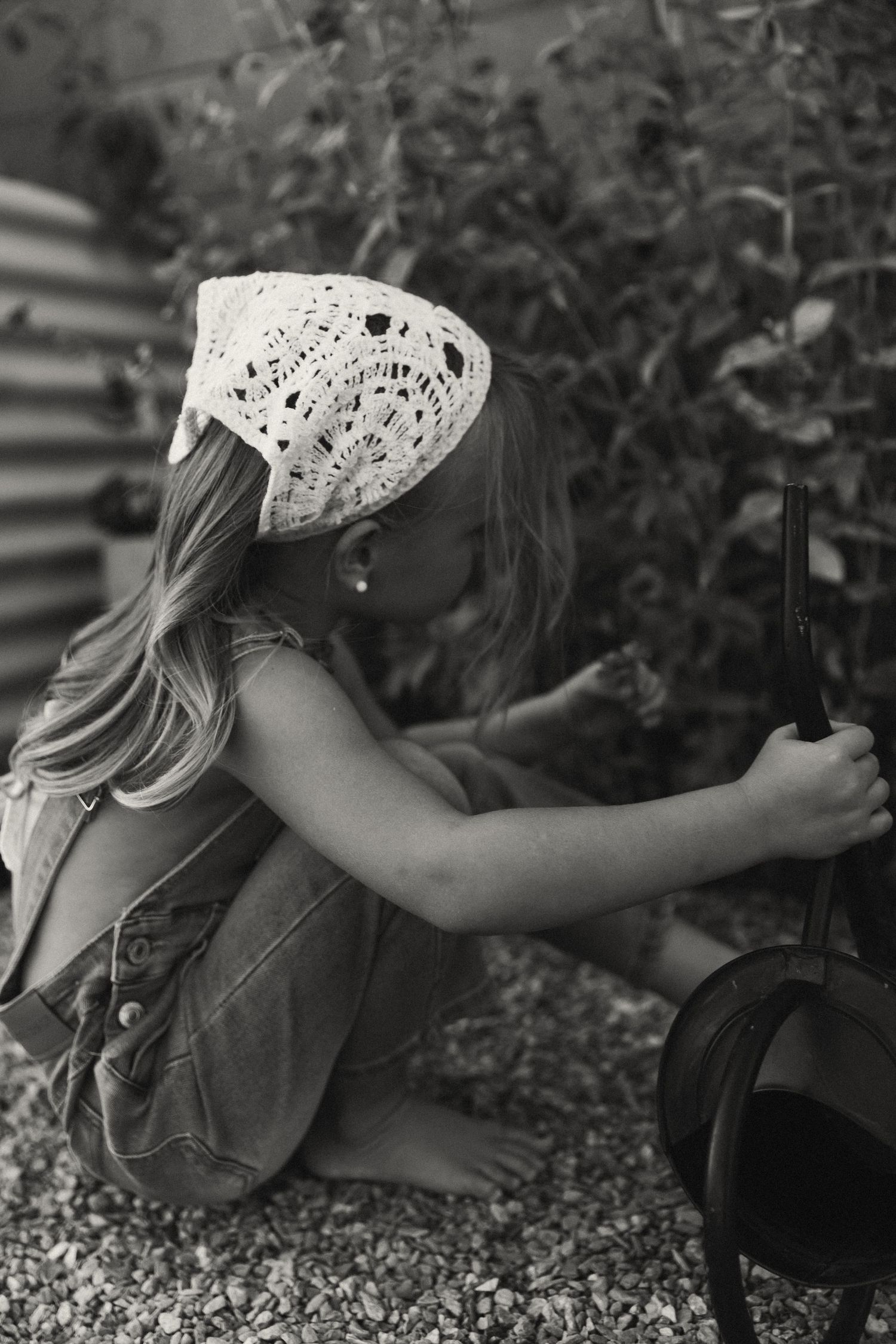 Black and white photo of a young barefoot girl in denim overalls and lace hat gardening outdoors.