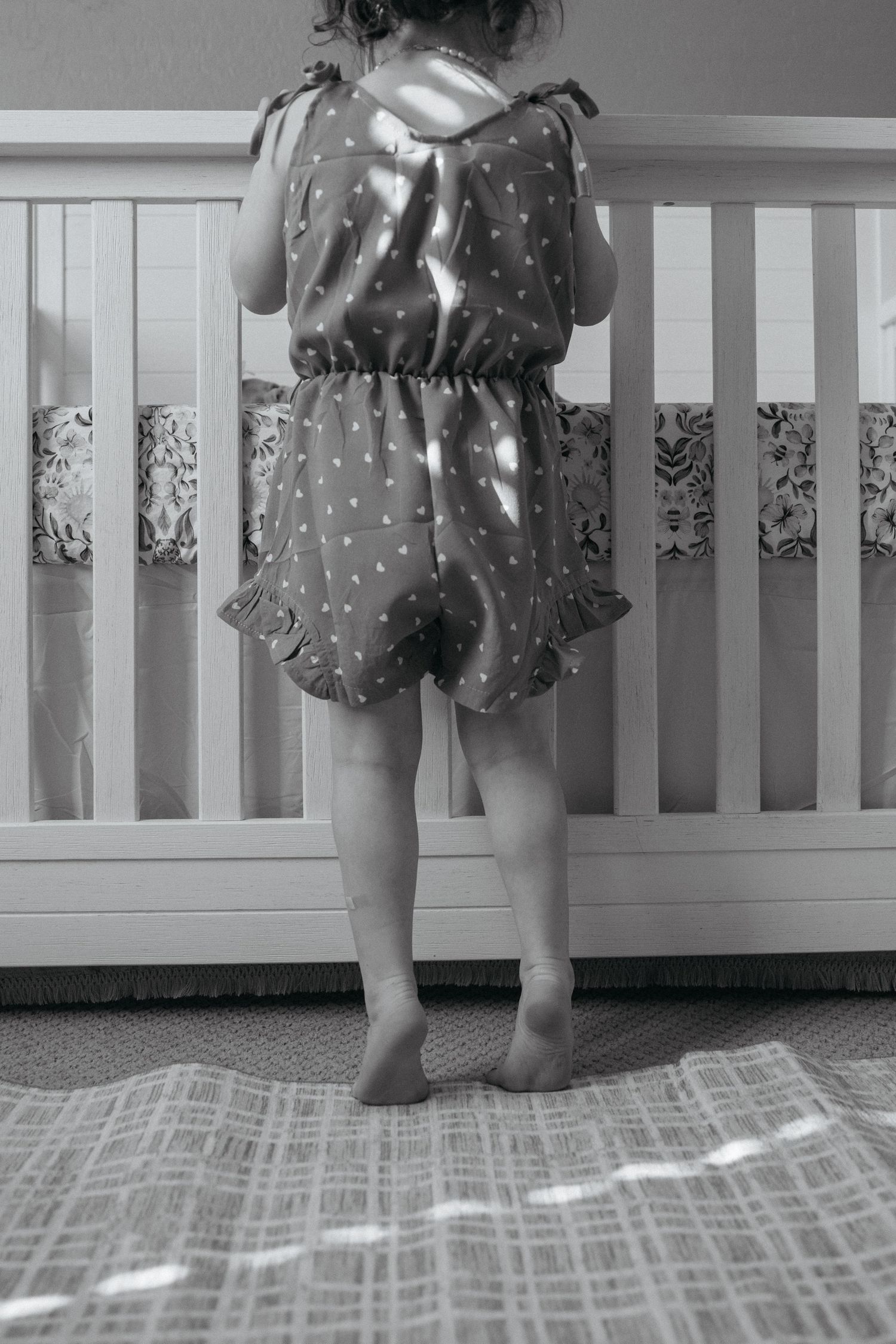 Black and white photo of a toddler girl in a polka dot dress standing at a white crib in a nursery.