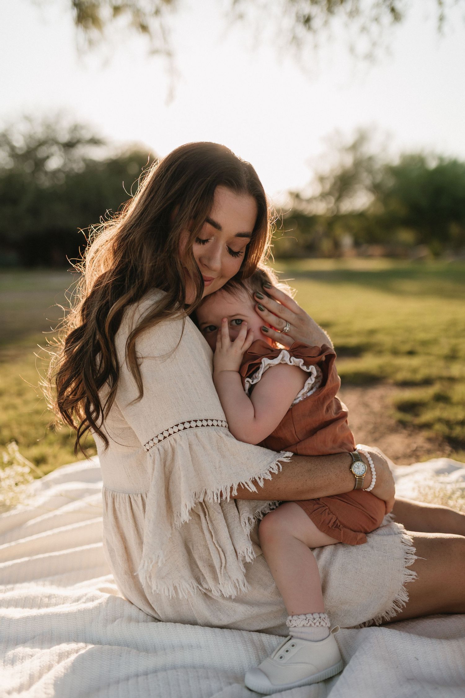 A parent in a white dress shares tender moments with a baby on a blanket in sunlit grass outdoors.