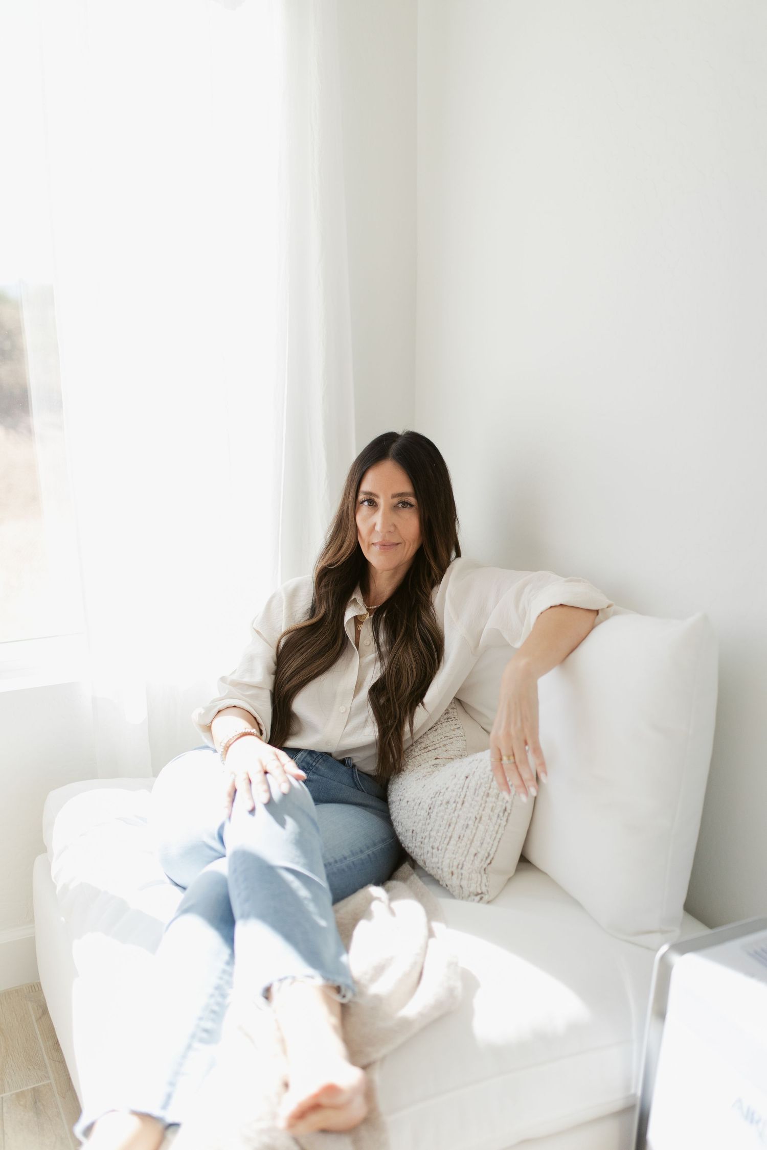 Woman in white sweater and jeans relaxing on white couch in minimalist bright room with natural lighting.