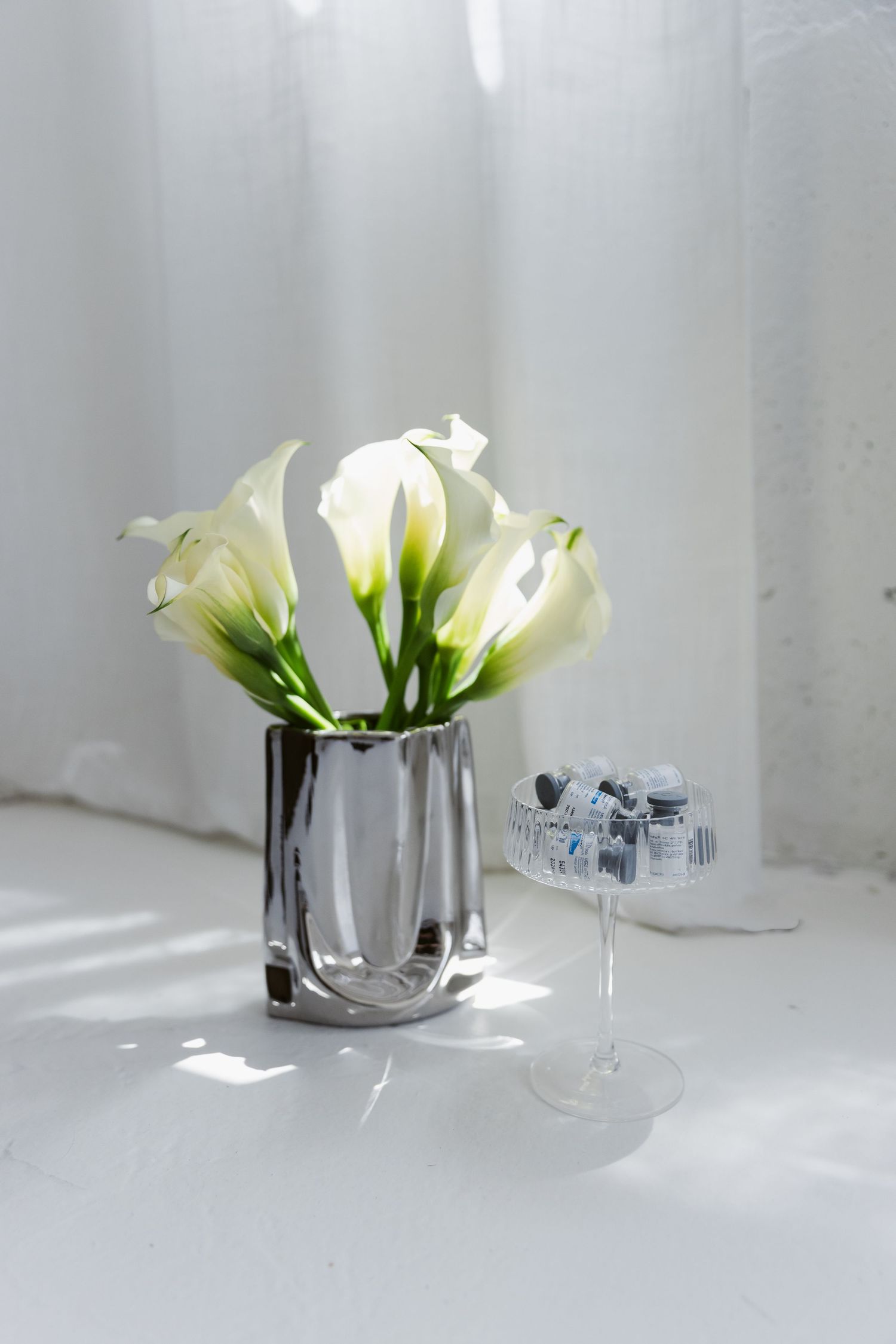 White calla lilies in a silver vase beside a crystal dish on a white surface with soft natural light.