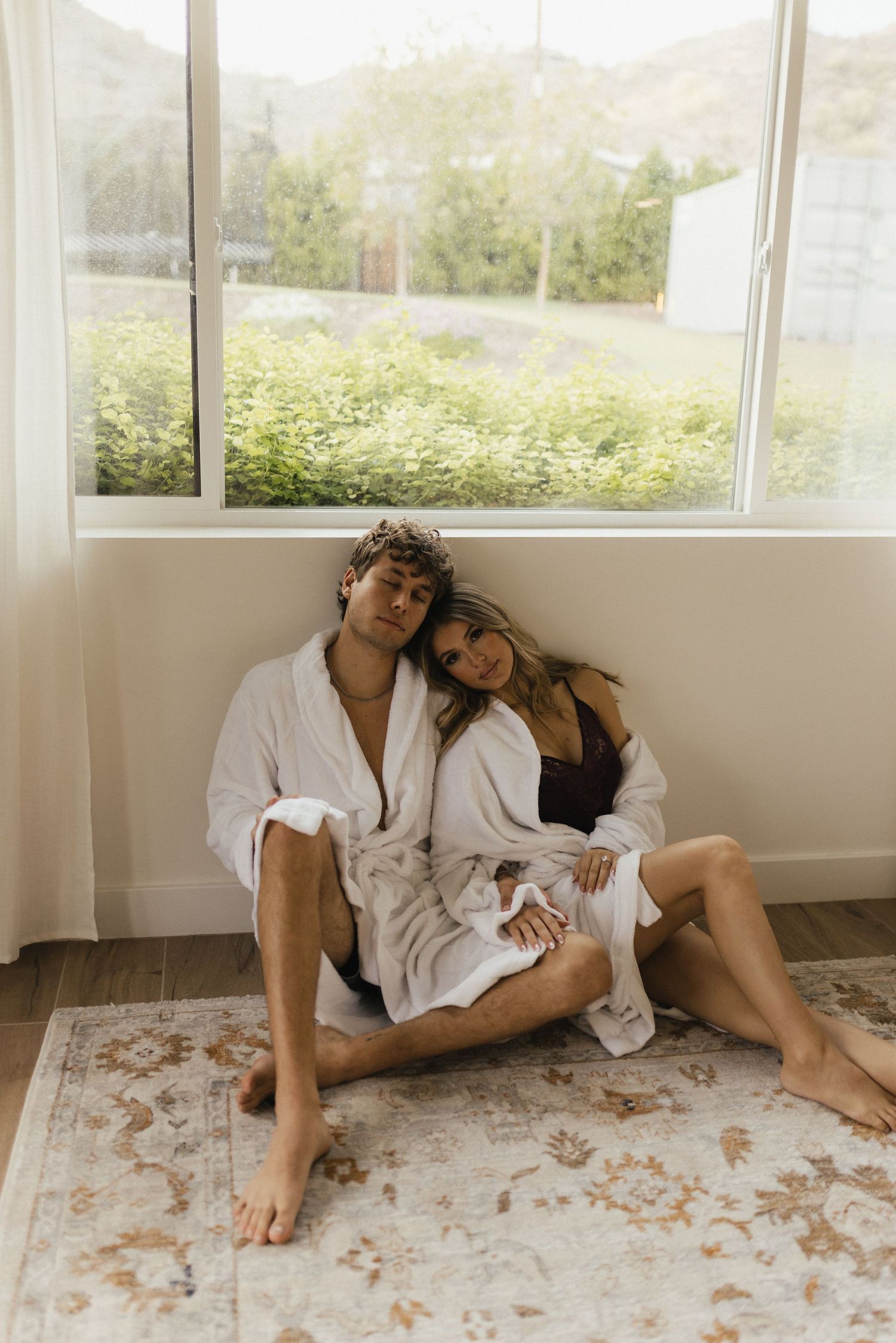 Couple in white robes sitting on a rug by a large window, leaning together in a cozy, relaxed pose.