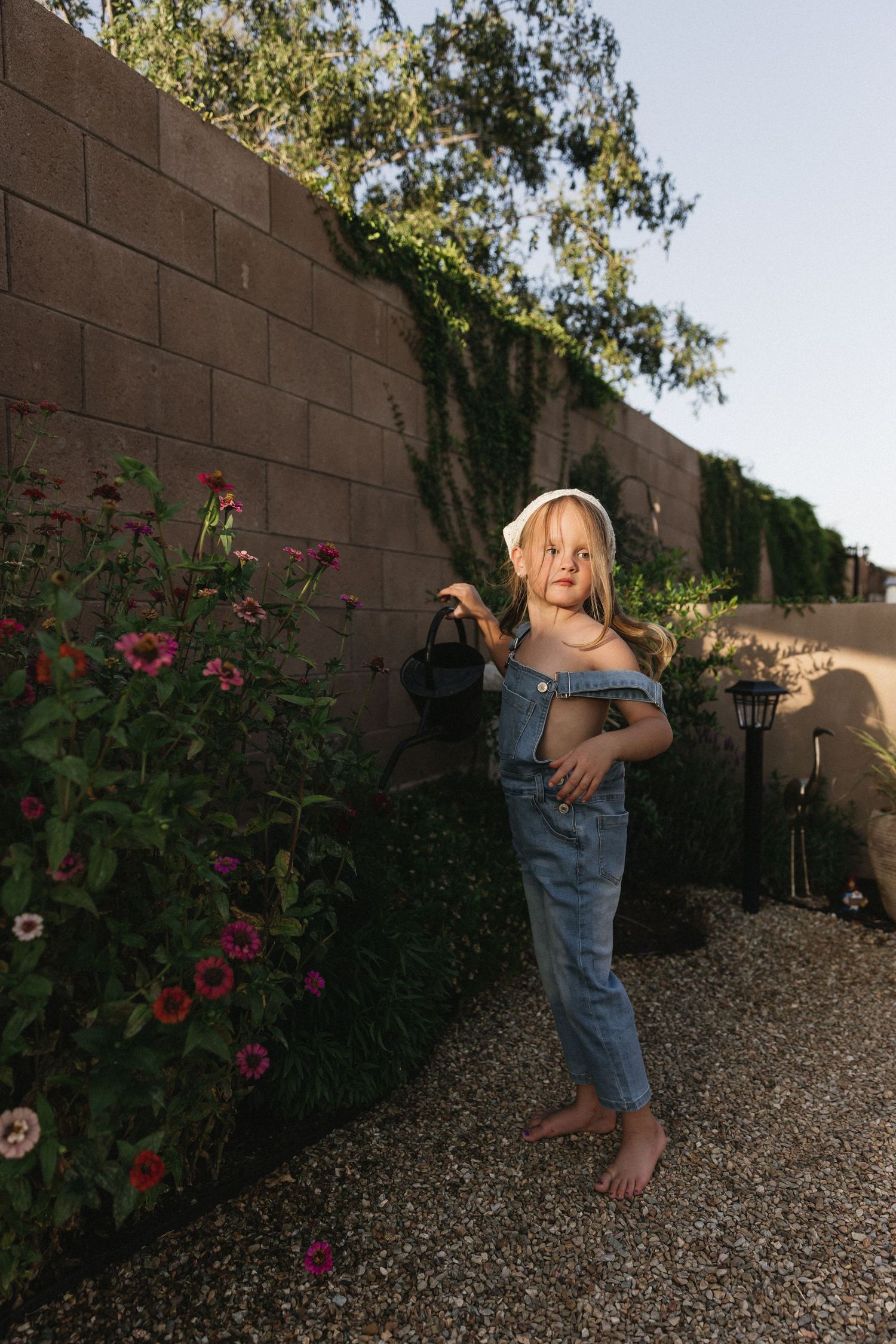 Young girl in denim overalls and white headband stands barefoot by a garden wall with pink flowers.