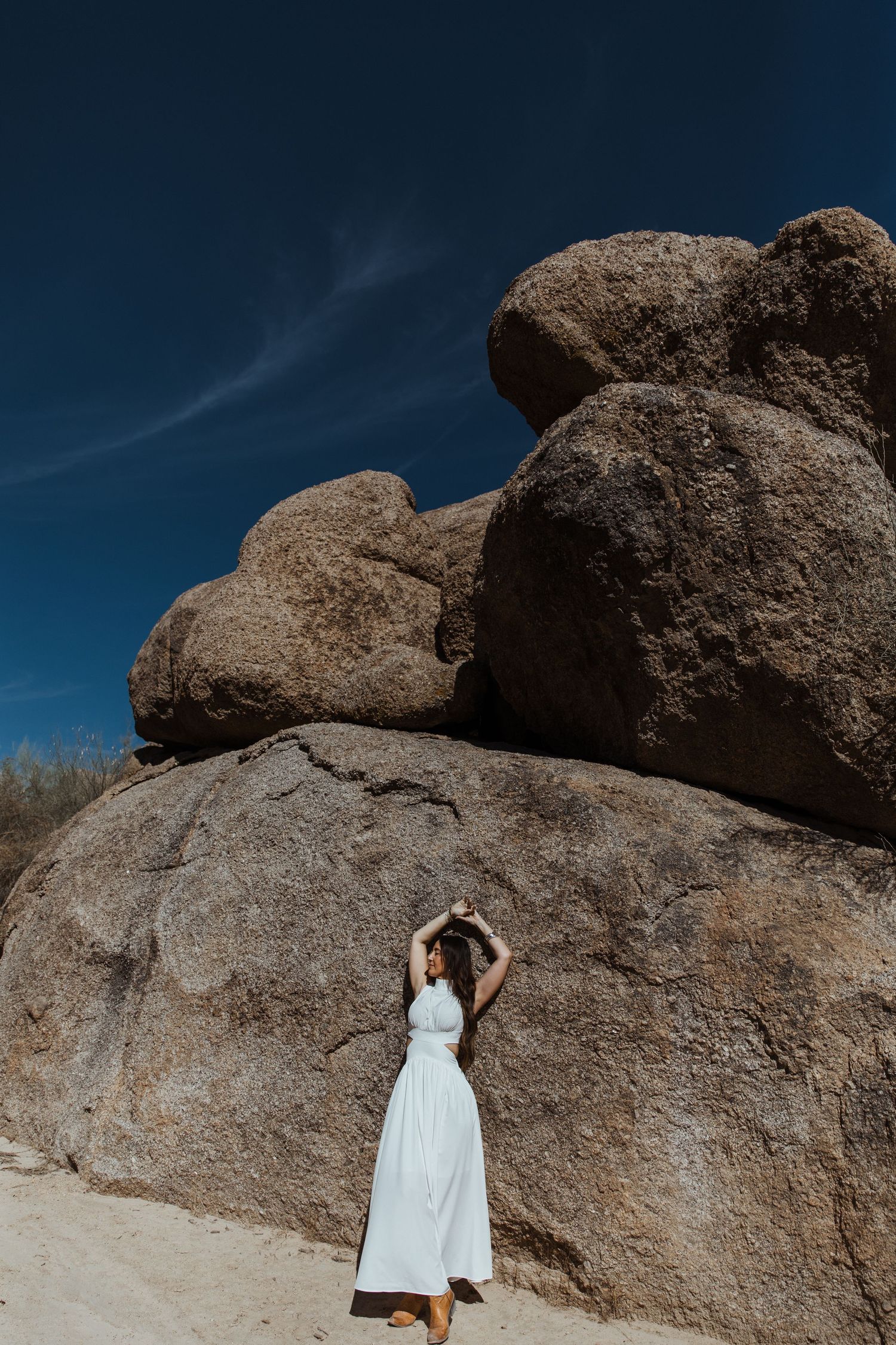 A person in a white dress poses gracefully against large desert boulders under a deep blue sky.