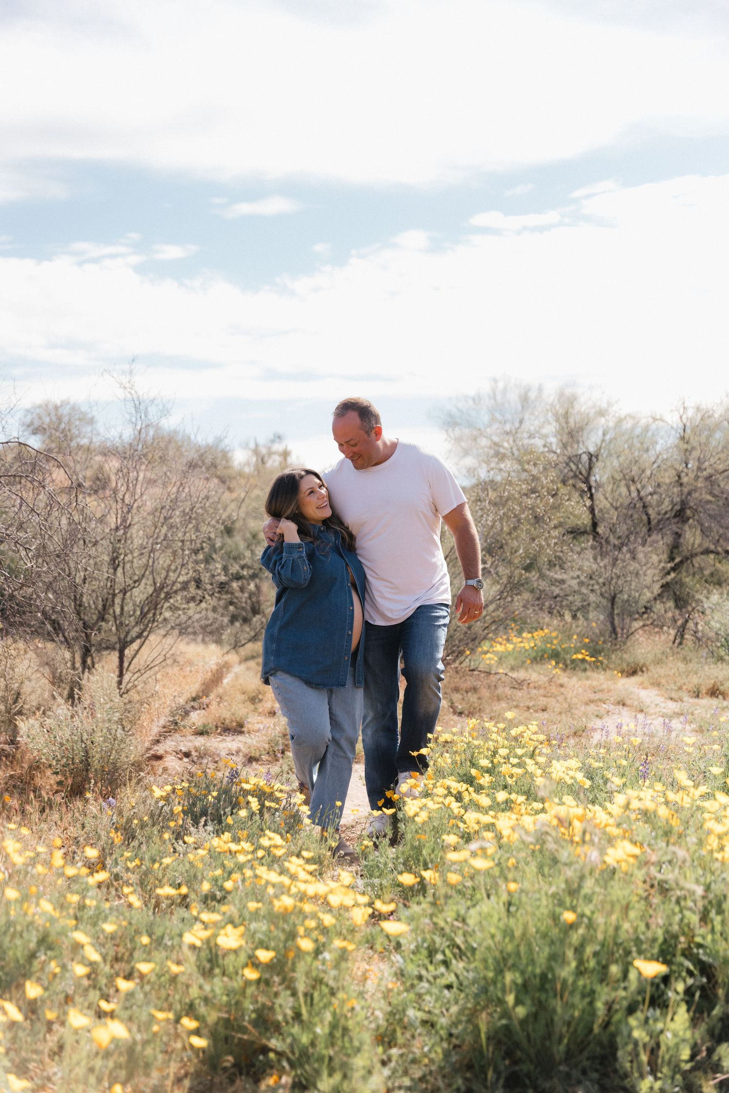 A couple walks together on a desert trail surrounded by yellow wildflowers and desert brush on a sunny day.