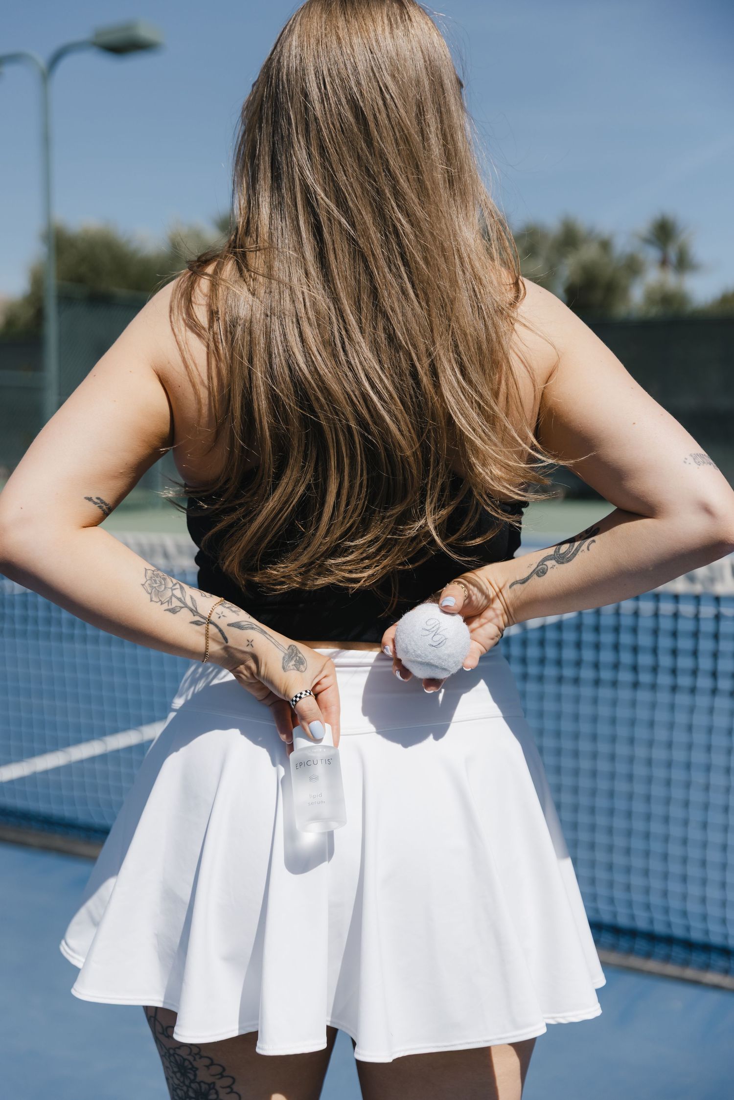 Woman in white tennis skirt and black crop top holding a tennis ball on a blue court, back view.