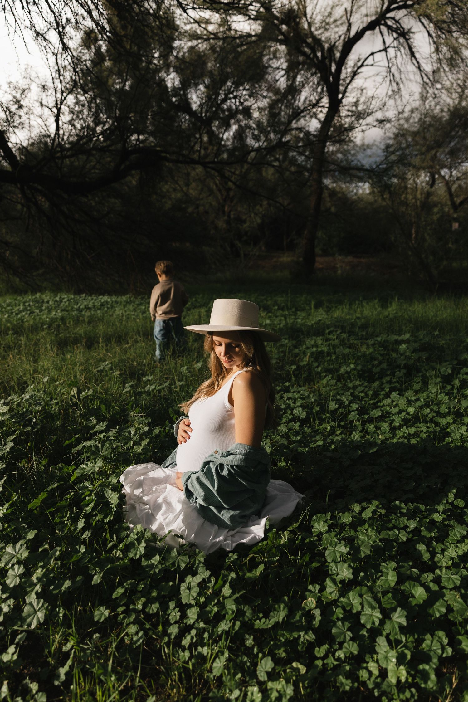 A figure in a white dress and wide-brimmed hat sits gracefully in a lush green field during golden hour.