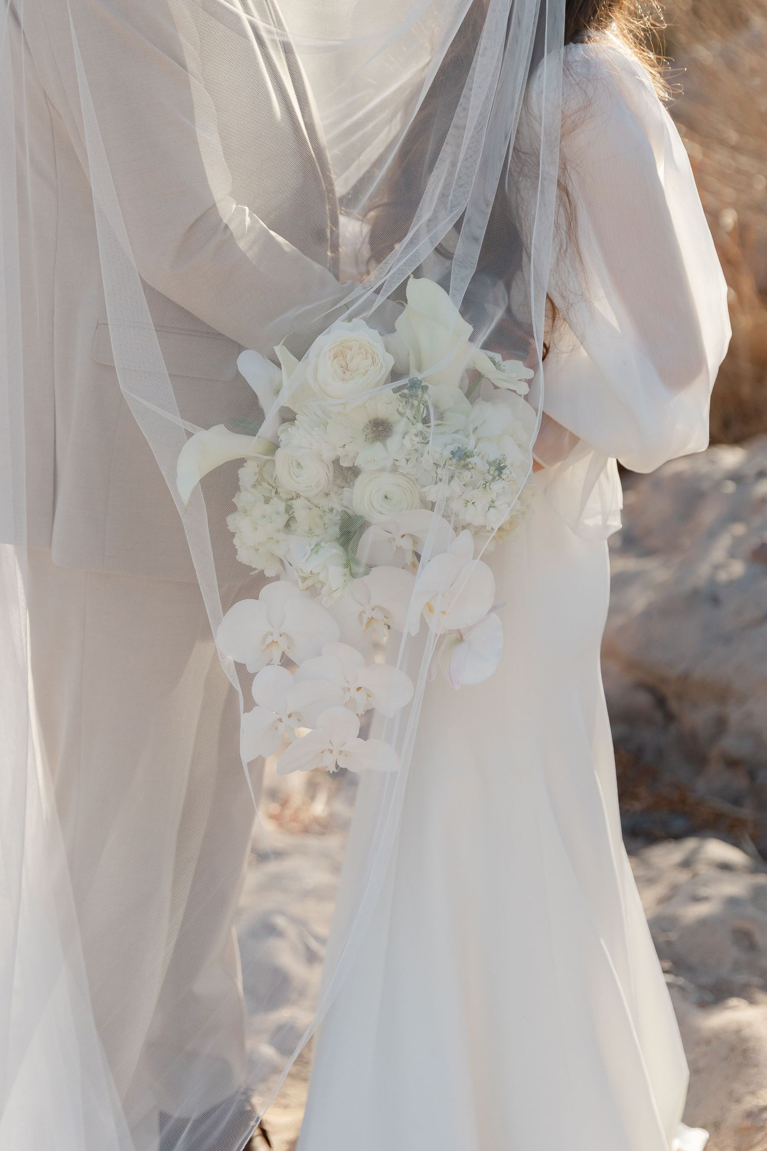 A delicate white bridal bouquet with roses and baby's breath against flowing sheer fabric and veil in soft sunlight.