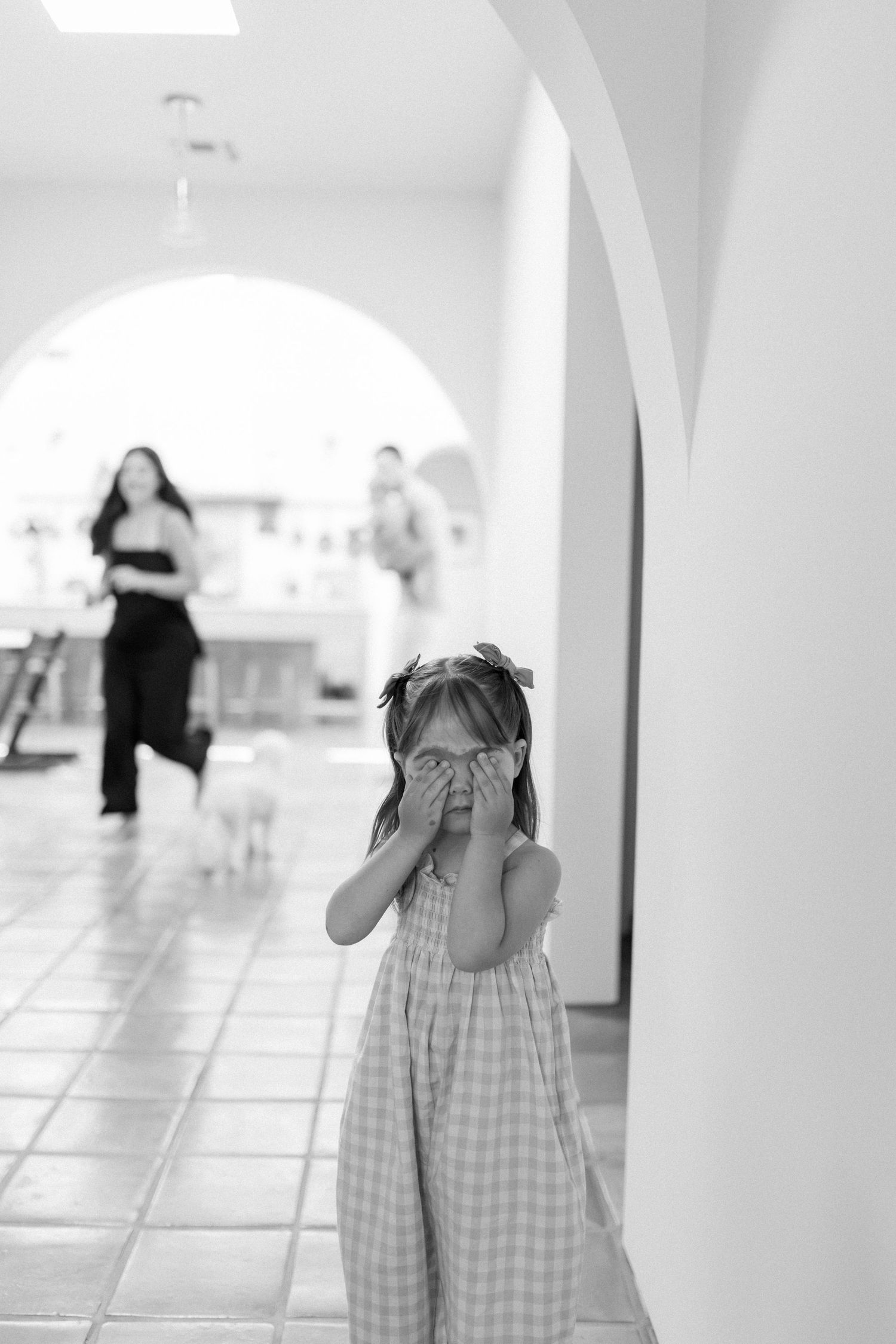 Black and white photo of a young girl in a plaid dress covering her face with her hands in a hallway.