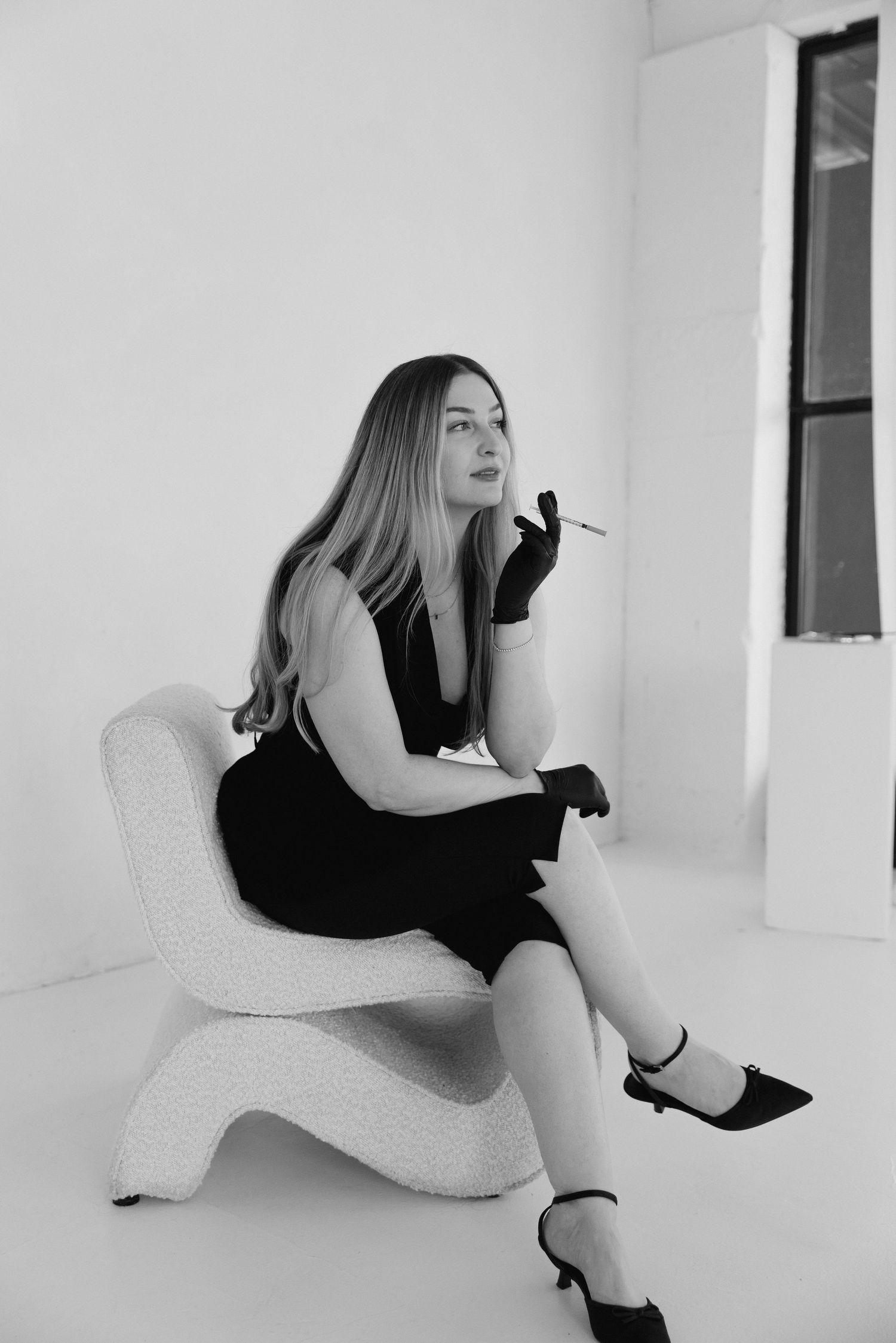 Black and white photo of a woman in a black dress and gloves sitting in a modern chair holding a cigarette.