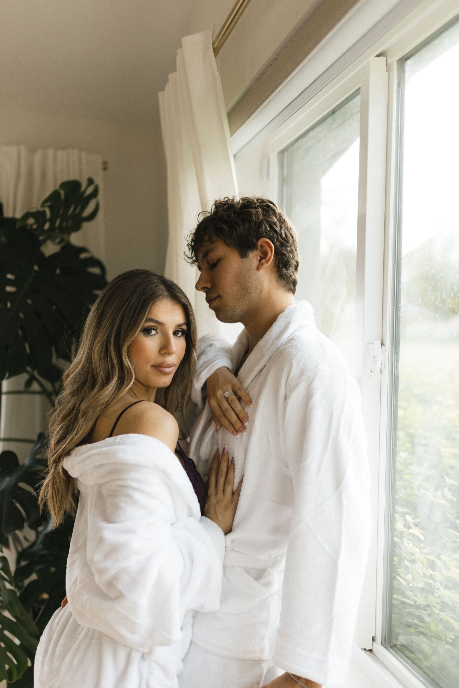 Couple in white robes embracing by a bright window with lush green plants in a cozy indoor setting.