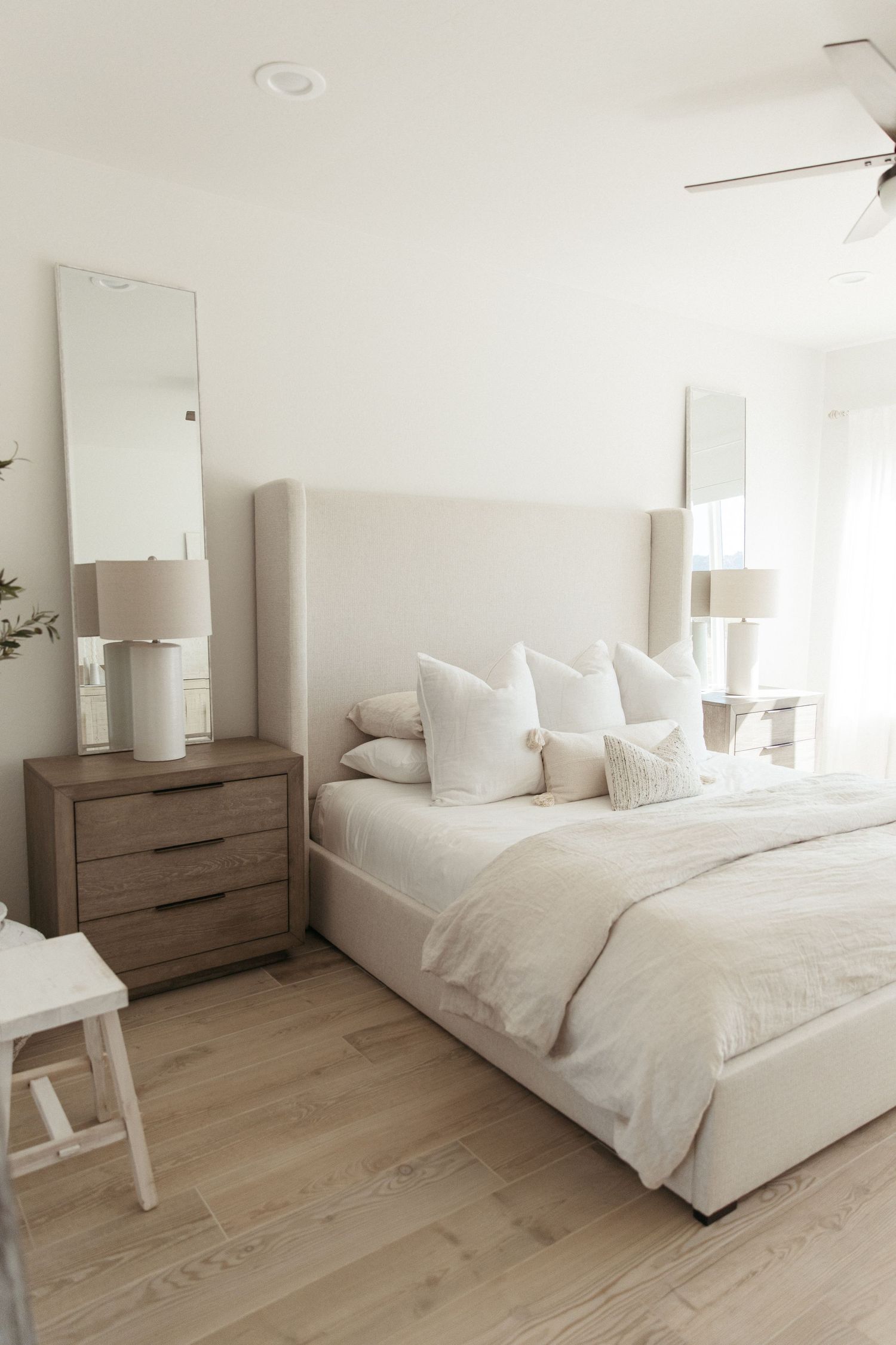 Modern minimalist bedroom with white bedding, light wood nightstand, and matching table lamps on neutral wood flooring.