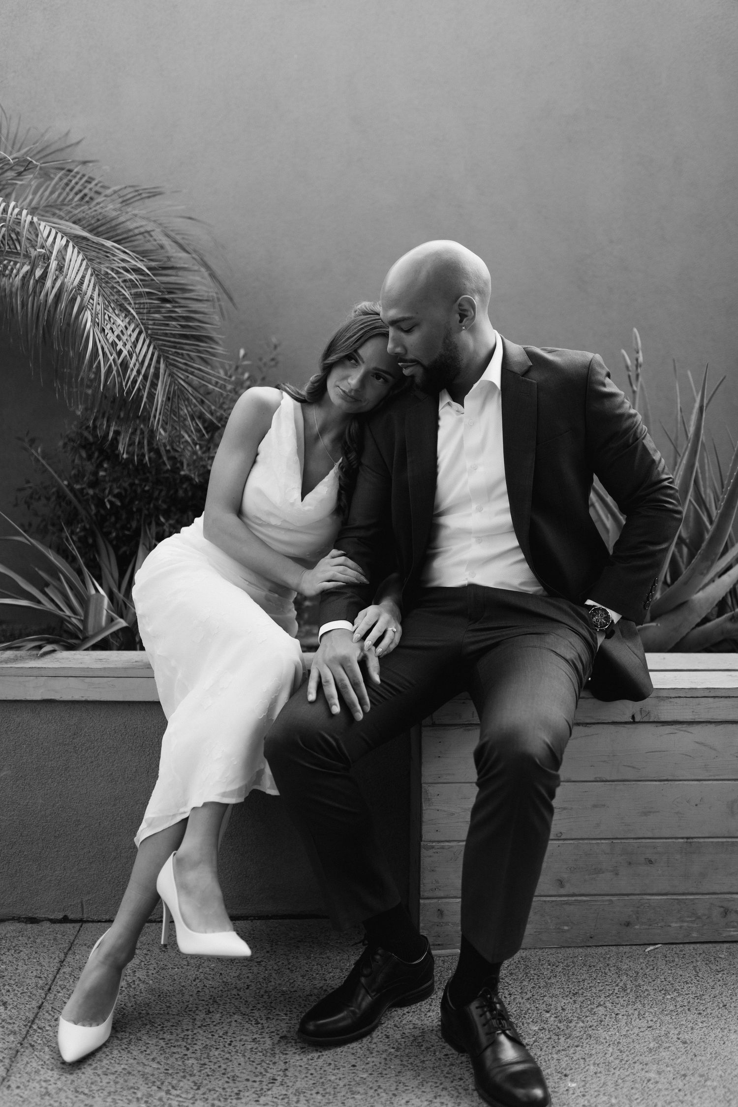 Romantic black and white portrait of couple in formal attire sitting close together on bench among palm fronds.