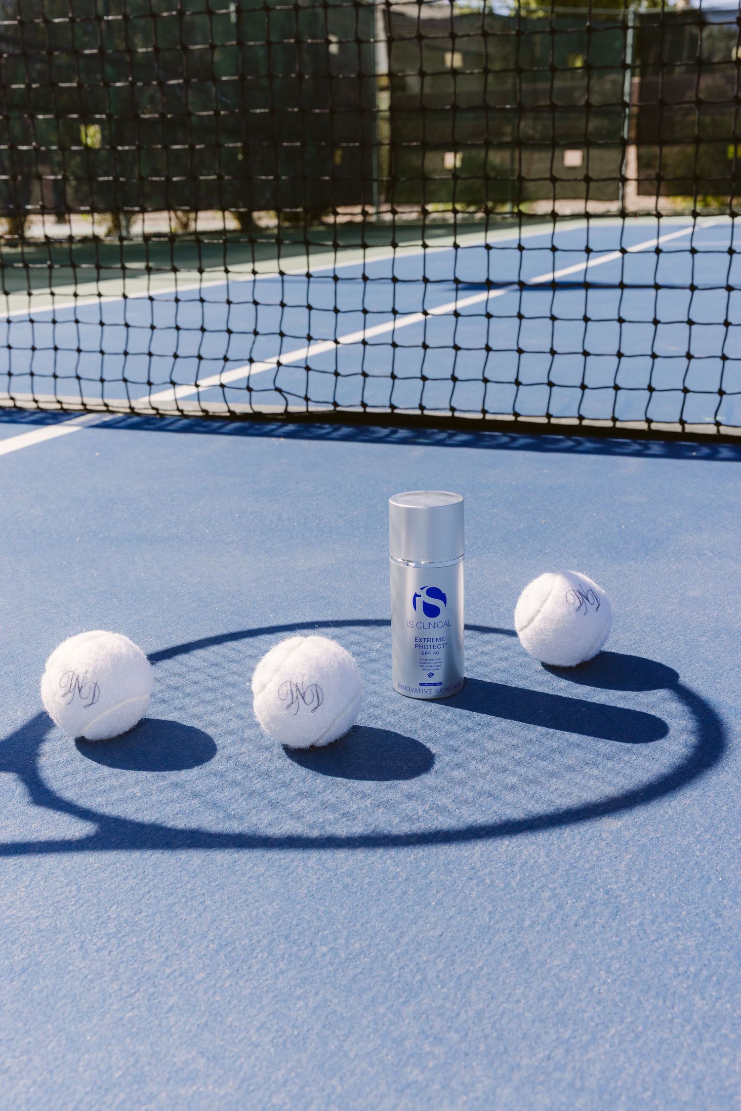 White tennis balls and iS Clinical skincare product arranged on a blue tennis court near the net.
