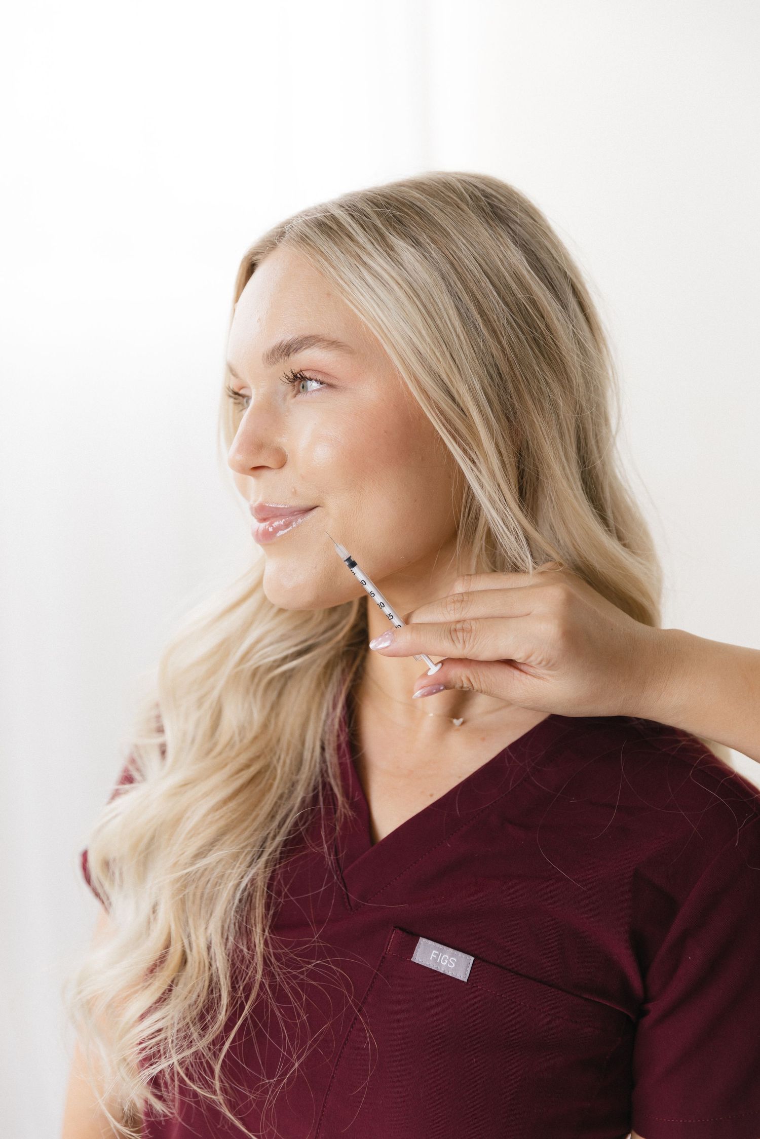 Blonde woman in burgundy scrubs having a necklace fastened around her neck, smiling softly.