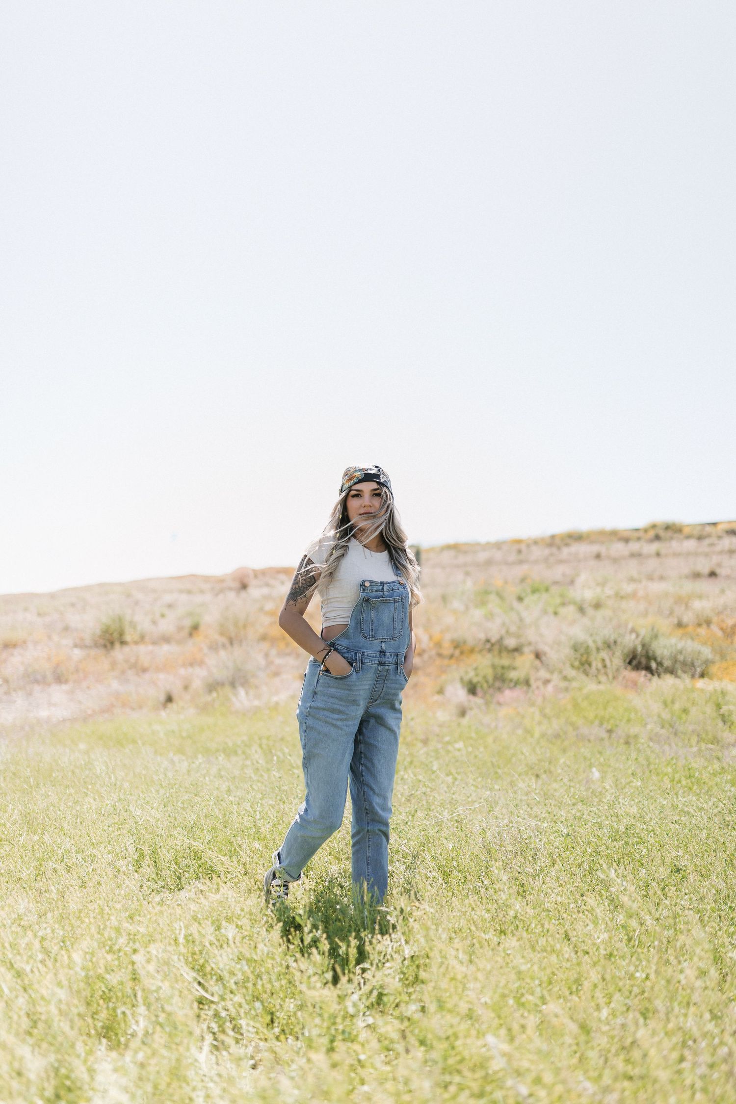 Person in denim overalls and sunglasses stands in a sunny meadow with dry grass and rolling hills in the background.