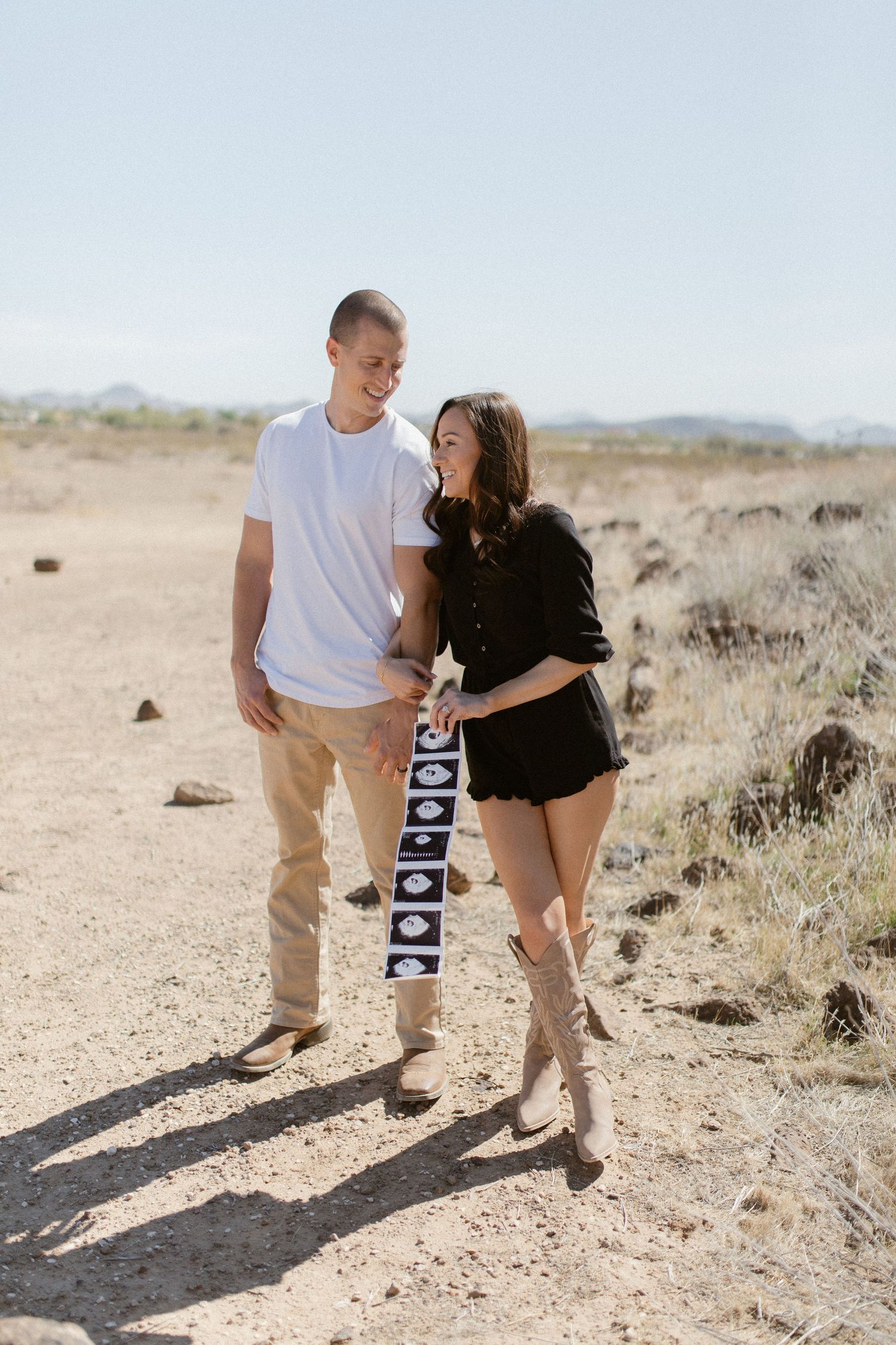 A couple stands together in a desert landscape holding ultrasound photos during a sunny pregnancy announcement photoshoot.