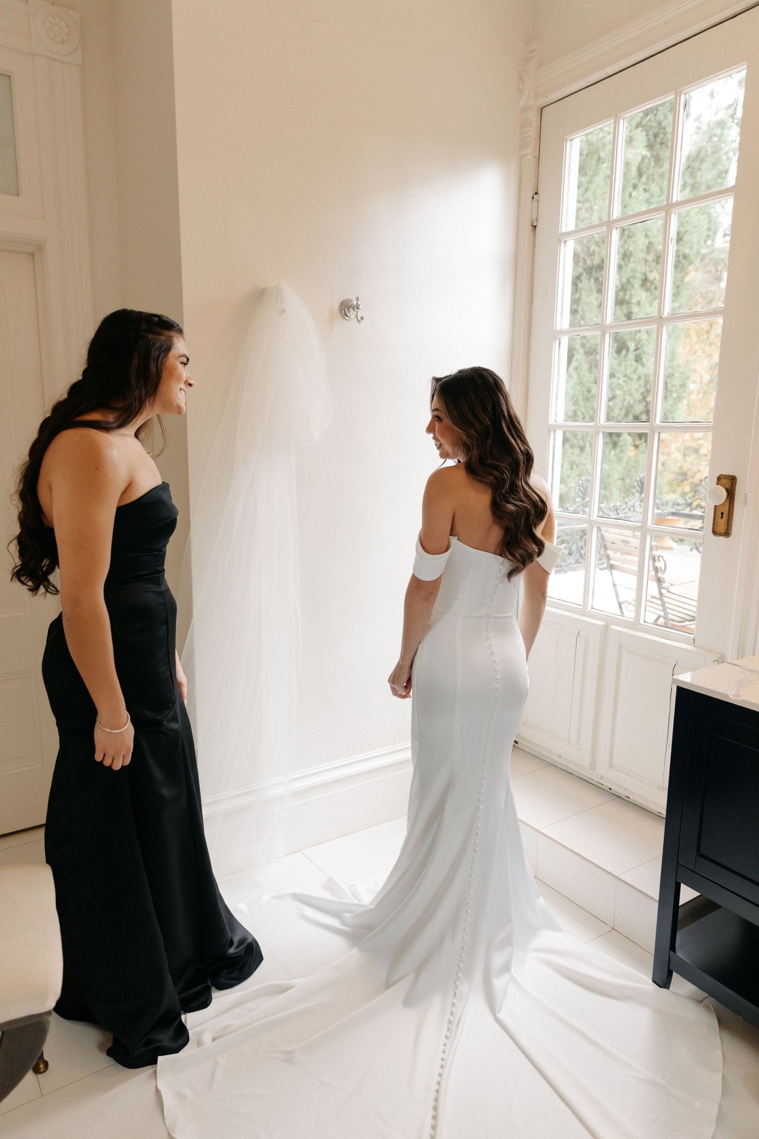 Two women in contrasting black and white dresses share a joyful moment in a bright sunlit room.