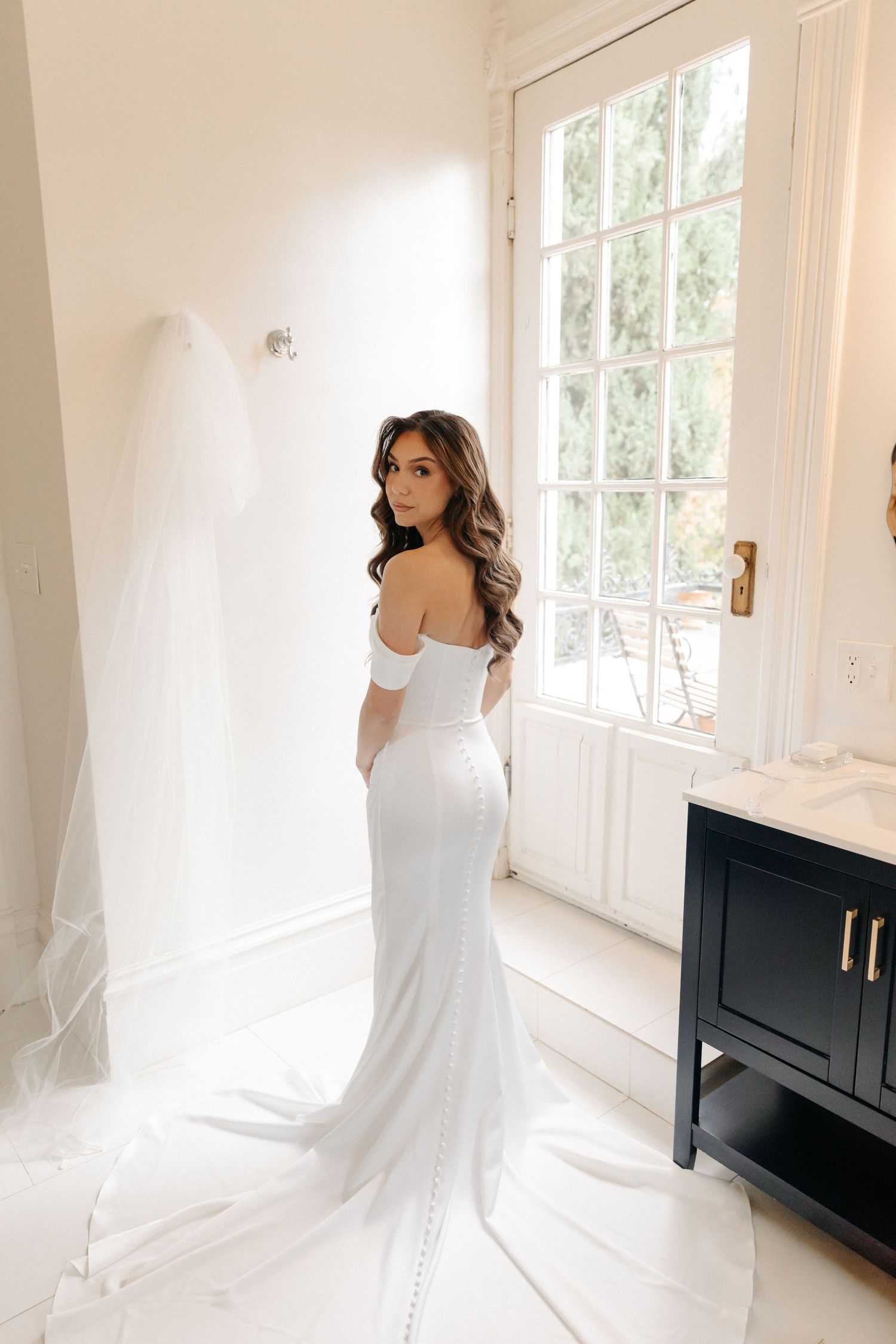 A bride in a white wedding dress poses elegantly by sunlit windows in a bright interior space.
