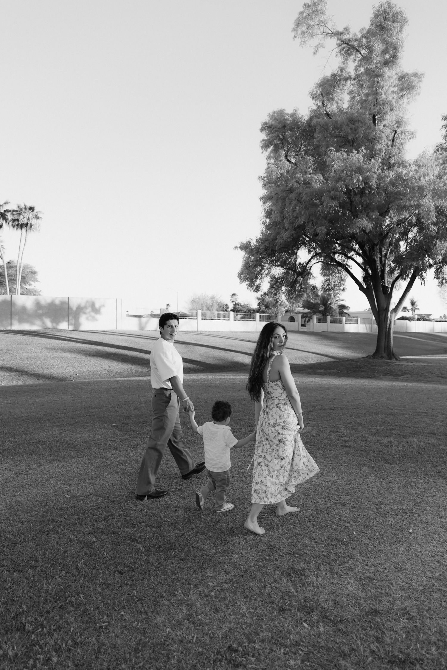 A family of three walks hand-in-hand across a grassy park in this black and white portrait photo.