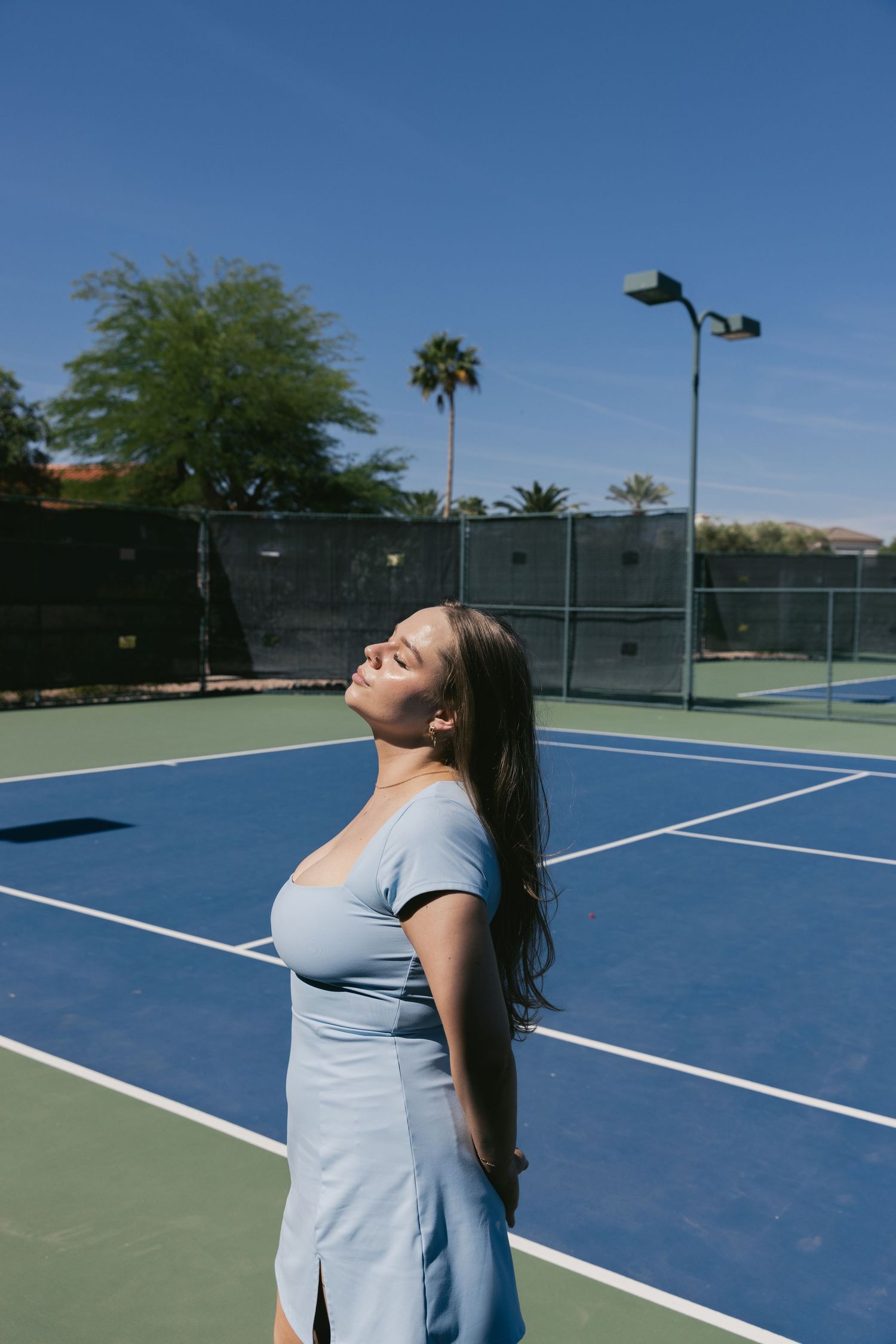 Woman in light blue dress posing on a blue tennis court with palm trees and clear sky in background.