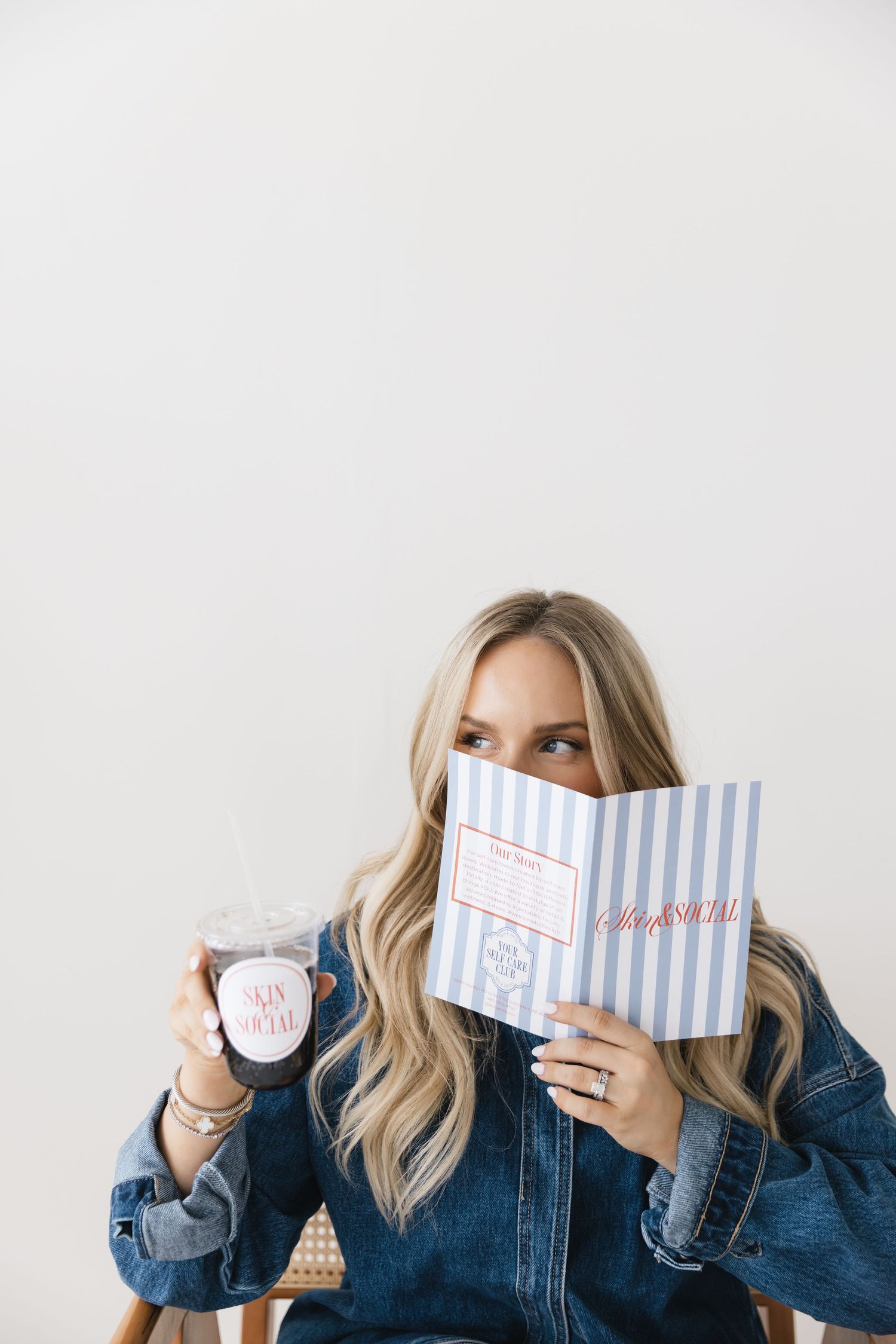 Blonde woman in denim jacket holding a striped book and pink drink cup while seated against a white background.