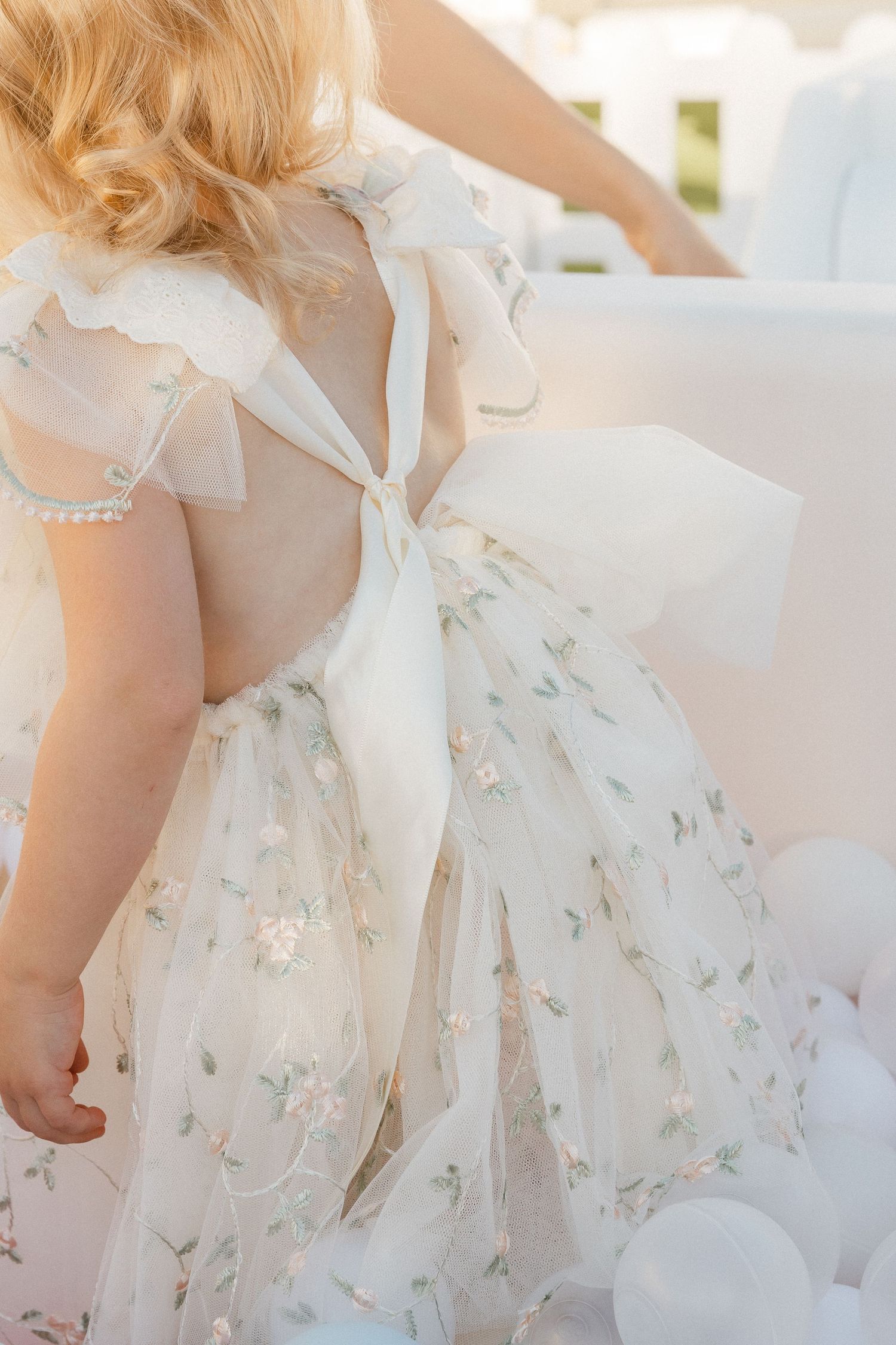 Delicate white floral dress with tulle and bow detail shown from behind against soft white balloons.