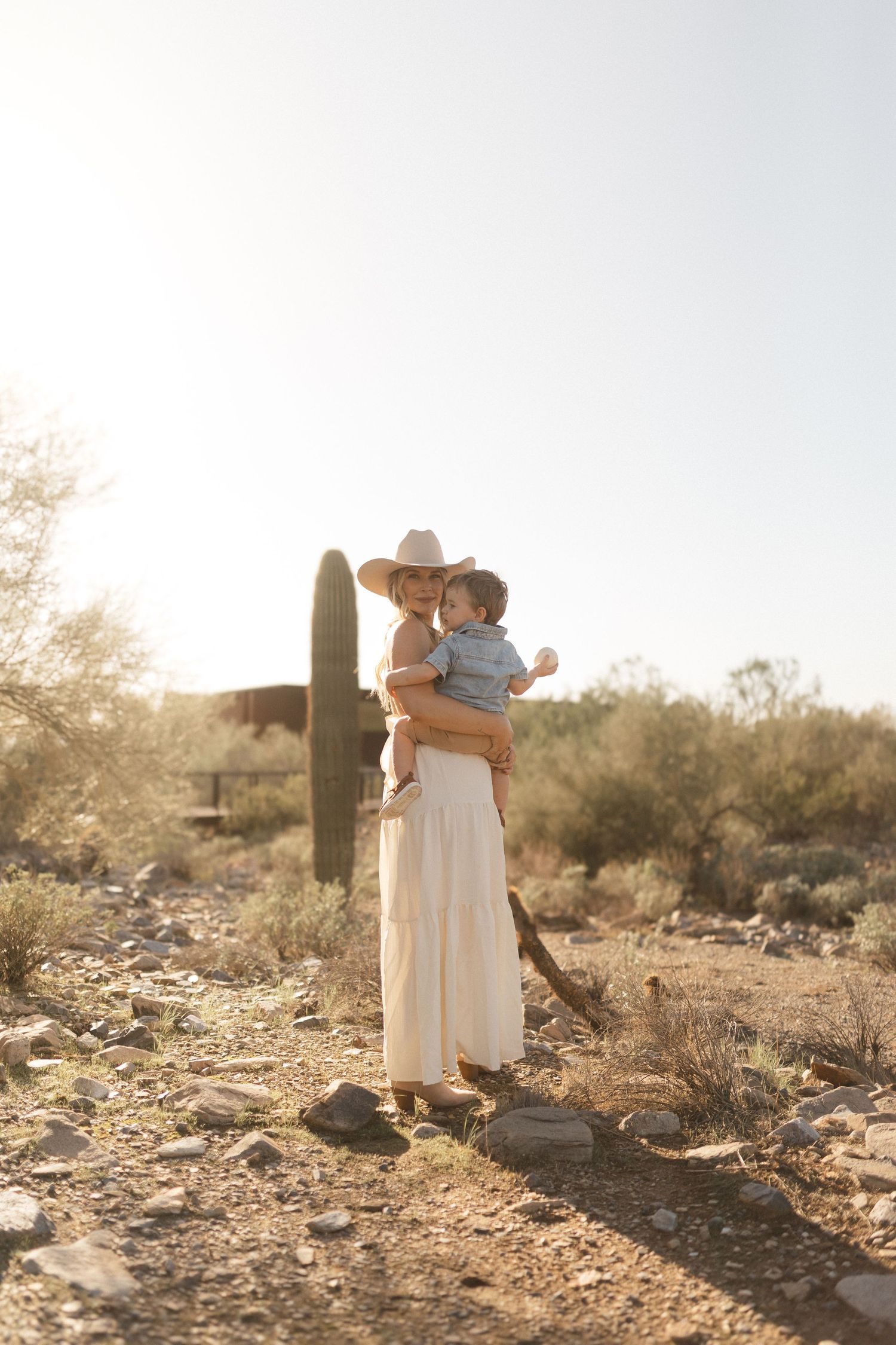 A figure in a flowing dress and hat stands in a desert landscape with saguaro cacti at golden hour.