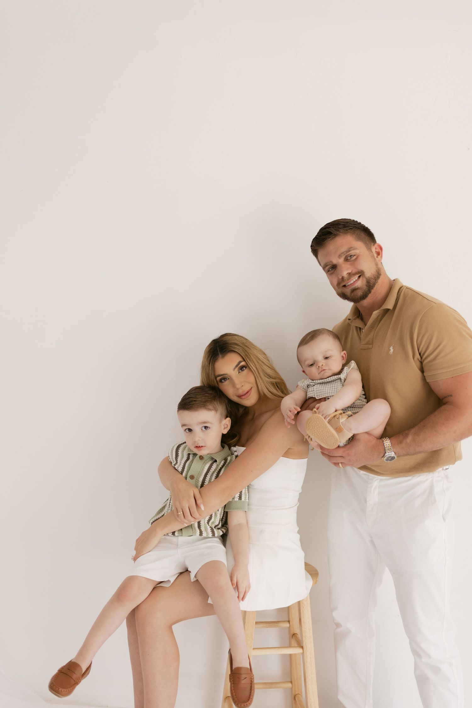 A smiling family of four poses together, with a woman holding a toddler and a man cradling a baby, against a white background.