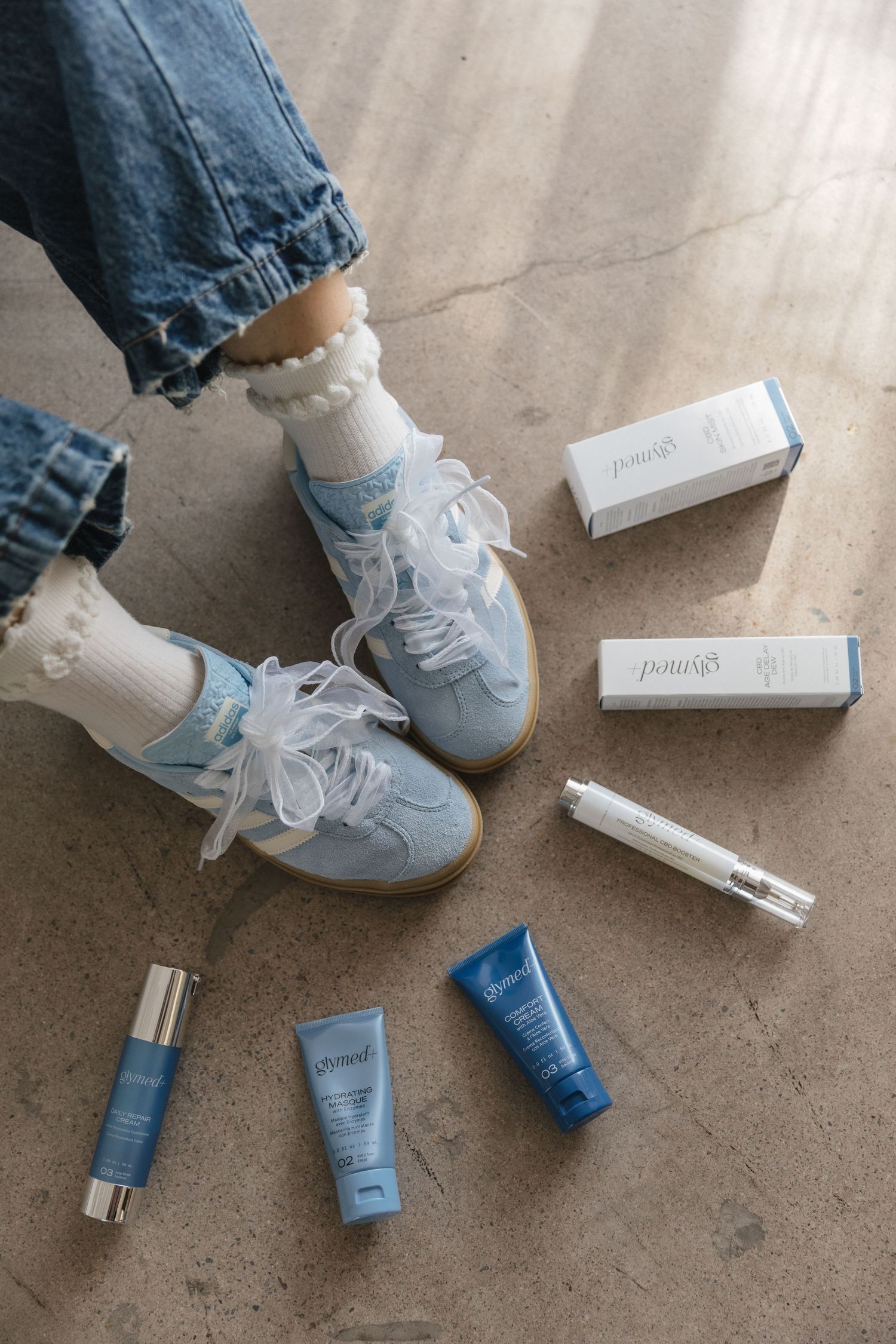 White sneakers with blue denim jeans and skincare products scattered on concrete floor showing a lifestyle product flatlay.