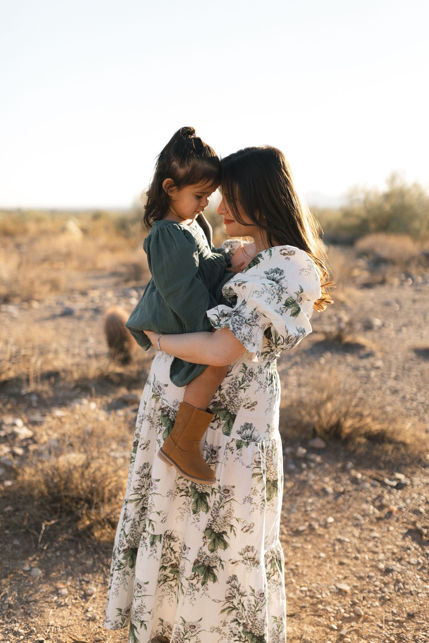 A parent and child embrace tenderly in filtered sunlight on arid desert terrain during a photo session.