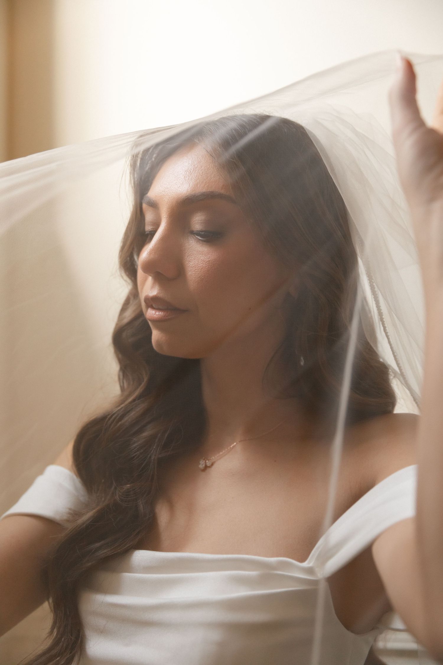 A bride in a white dress and veil poses with closed eyes in soft natural lighting.
