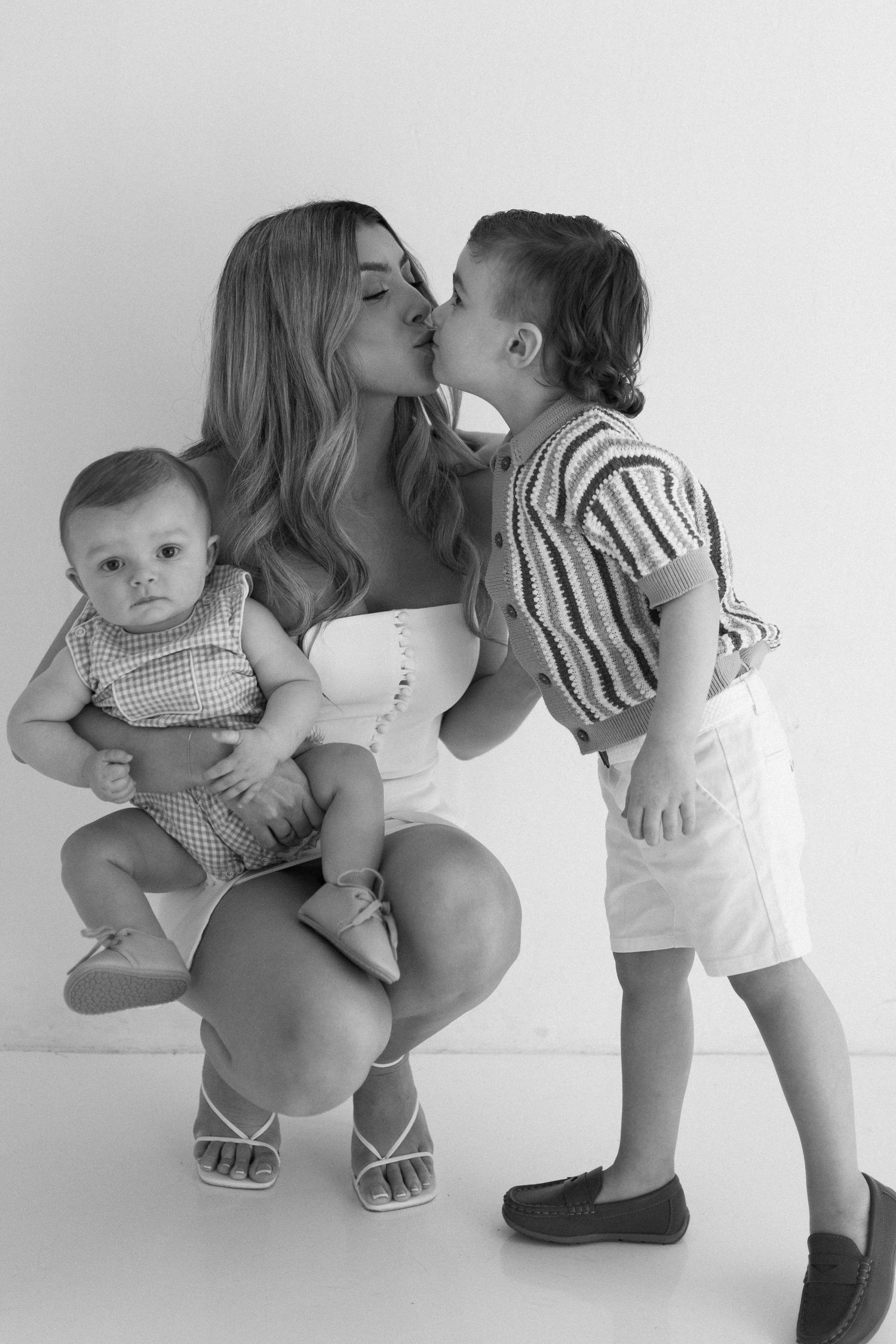 Black and white photo of a young mother crouching, holding a baby while an older child kisses her cheek.