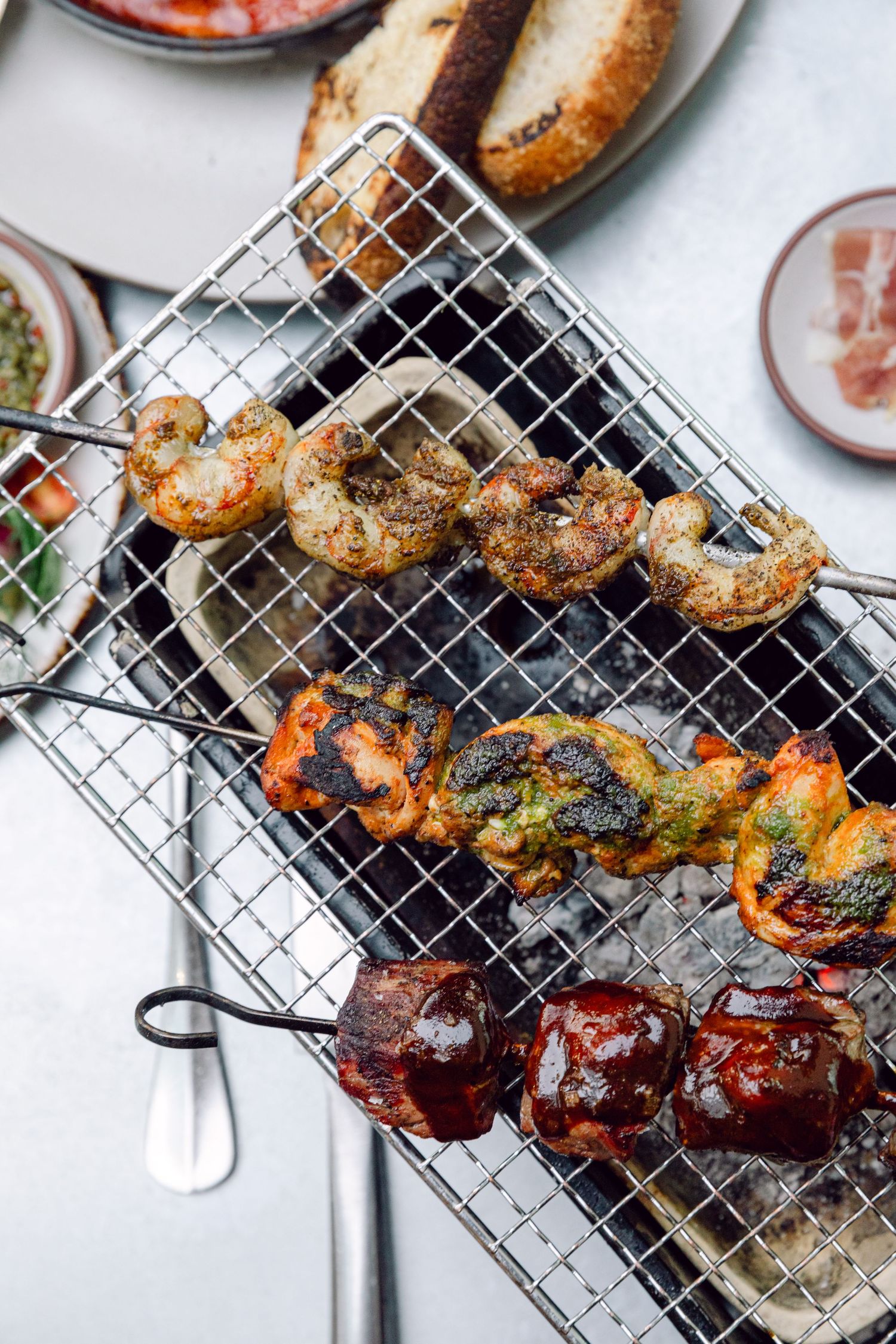 Grilled skewers of shrimp and vegetables on a wire cooking rack with various dipping sauces on the side.