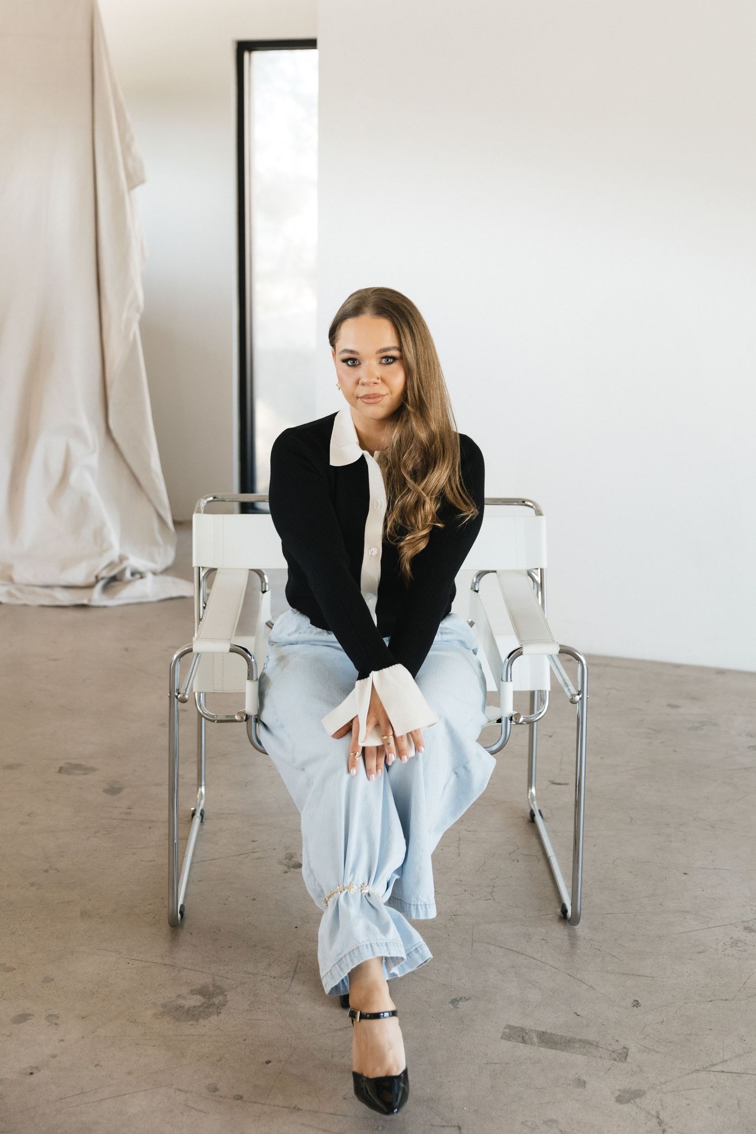 A person in a black blazer and light jeans sits casually in a modern white chair against a minimalist background.