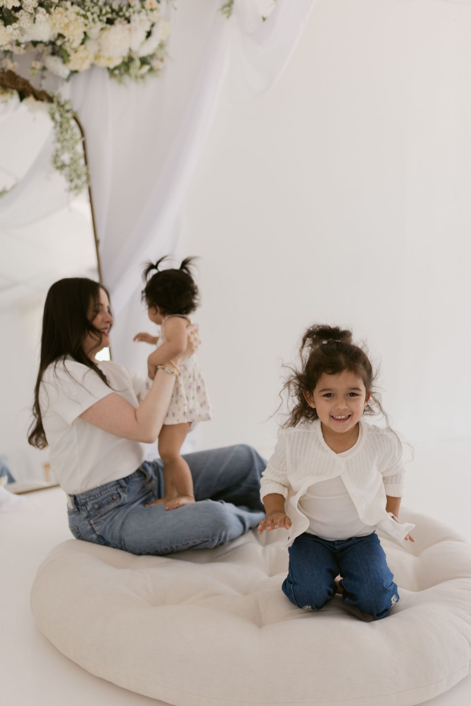 A woman sits on a white cushion with two young girls in a bright, floral-decorated studio setting.