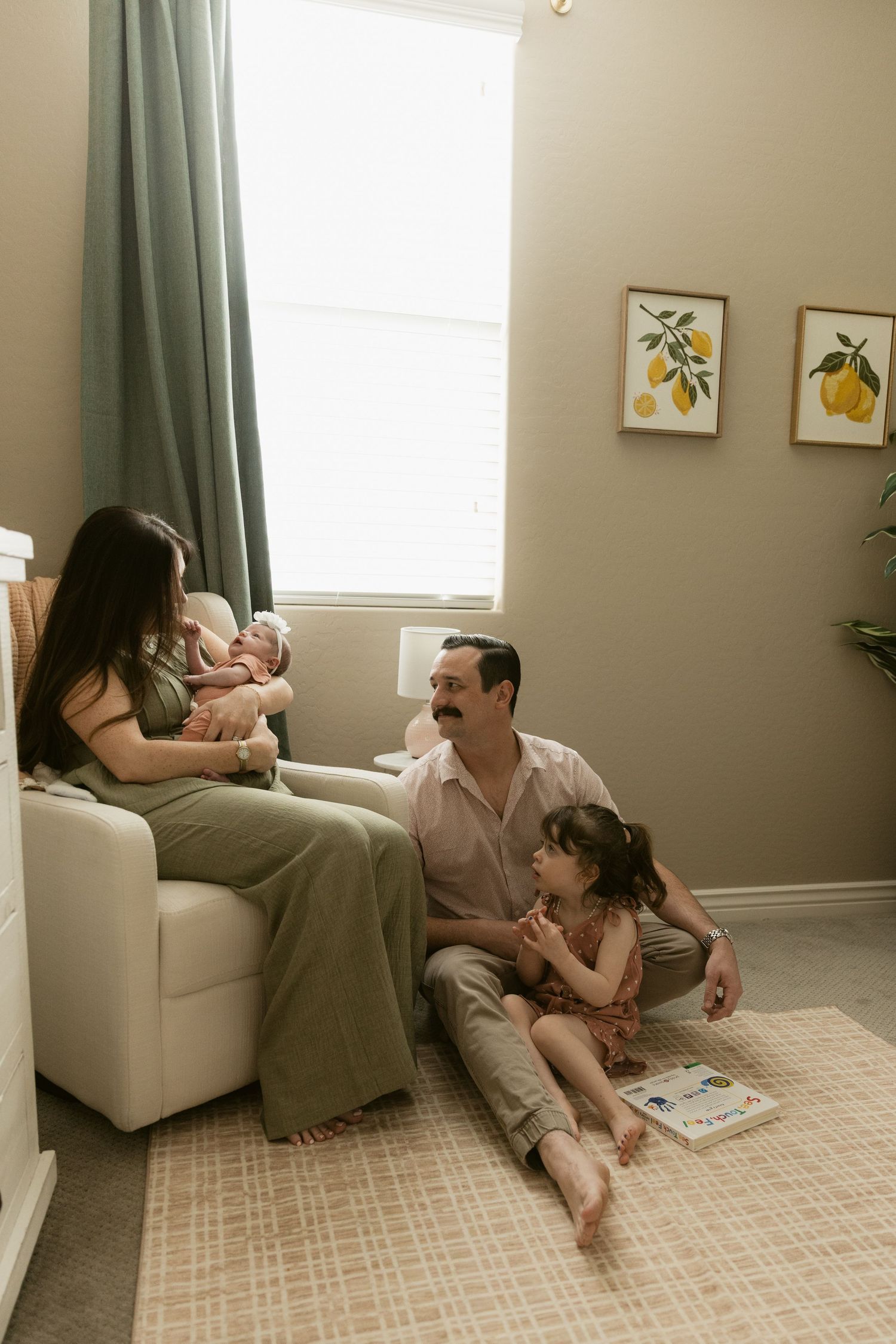 A family relaxes in a nursery, mom holding newborn in chair while dad sits on floor with toddler daughter.