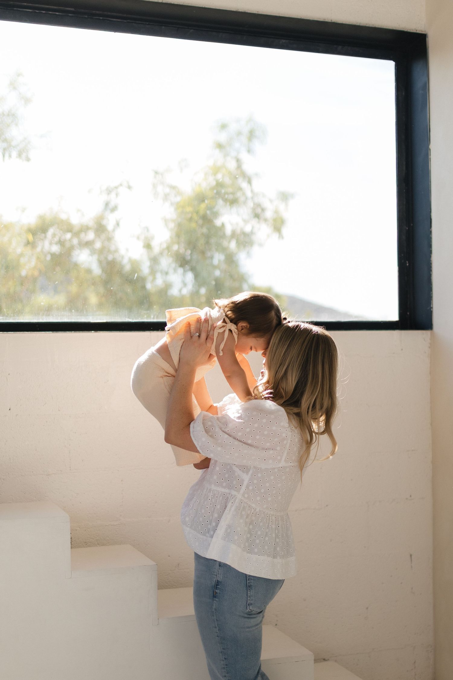A person in blue jeans and white shirt leans over to look out a modern window in a minimalist white room.