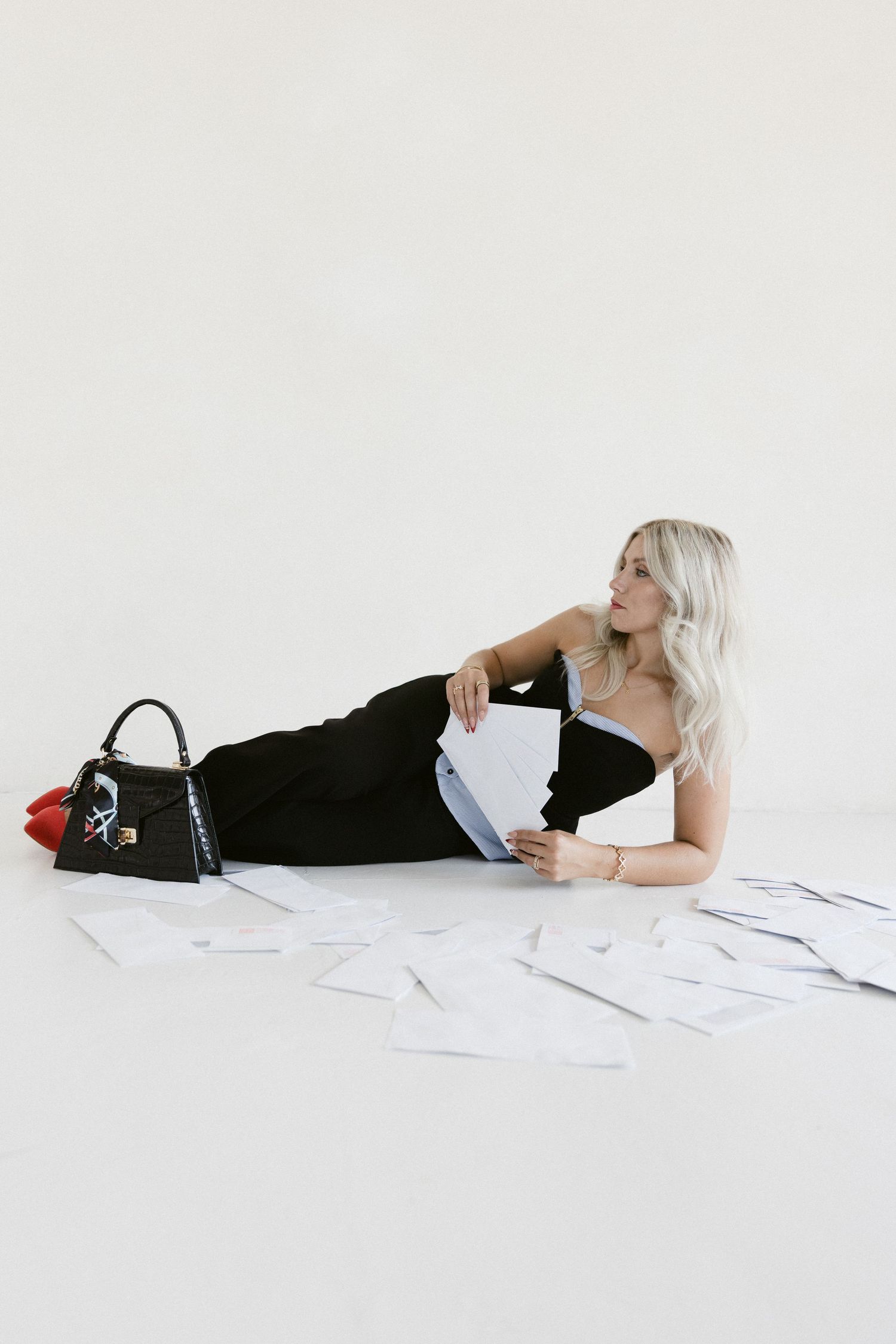 Blonde woman in black dress lying on white floor surrounded by papers with a black handbag beside her.