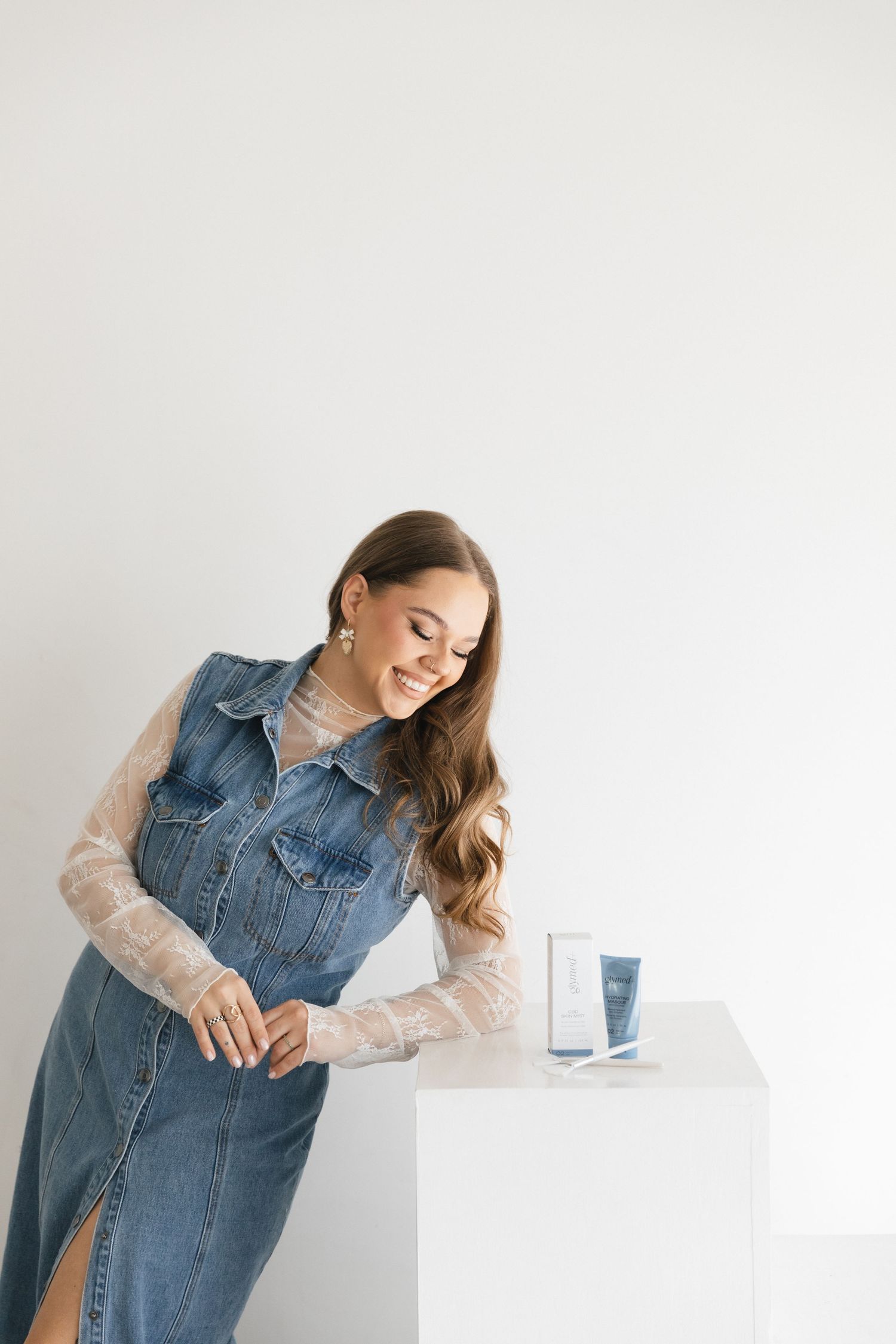 Woman in denim vest and sheer sleeves smiles while holding a glass of water against white background.