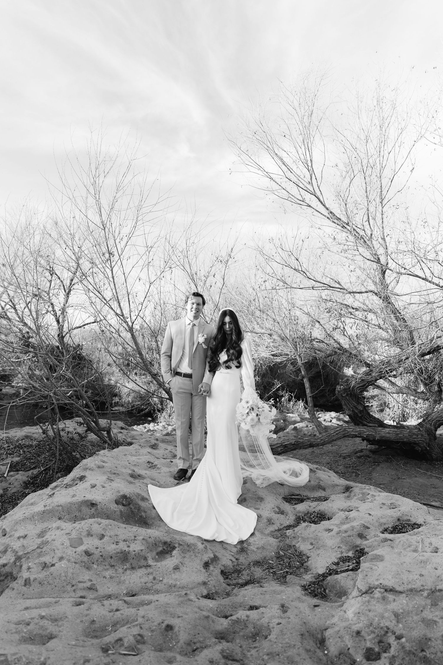 A couple stands on rocky terrain surrounded by bare winter trees in a black and white outdoor photo.
