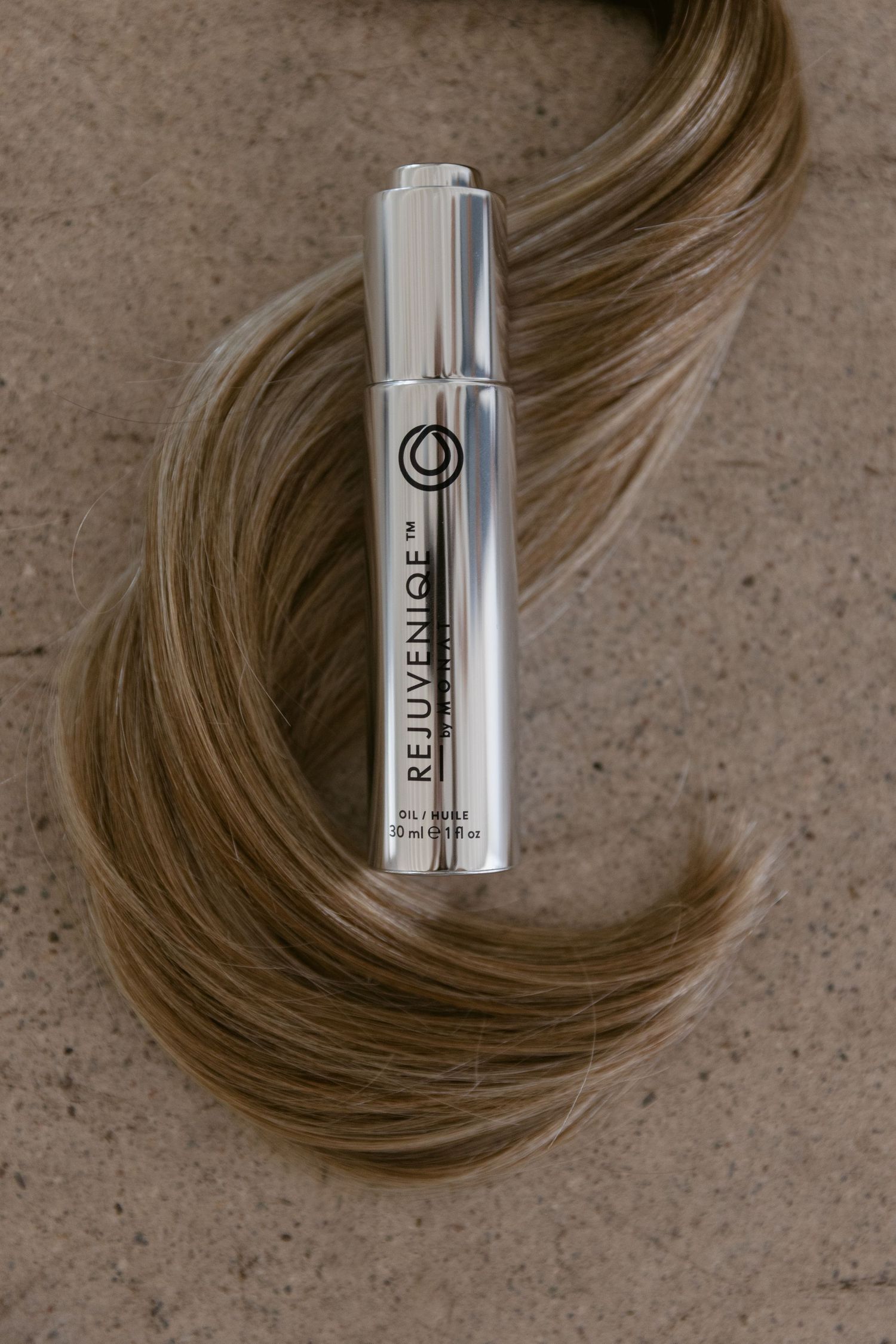Rejuveniqe hair oil serum in silver bottle resting on silky blonde highlighted hair strands on stone surface.