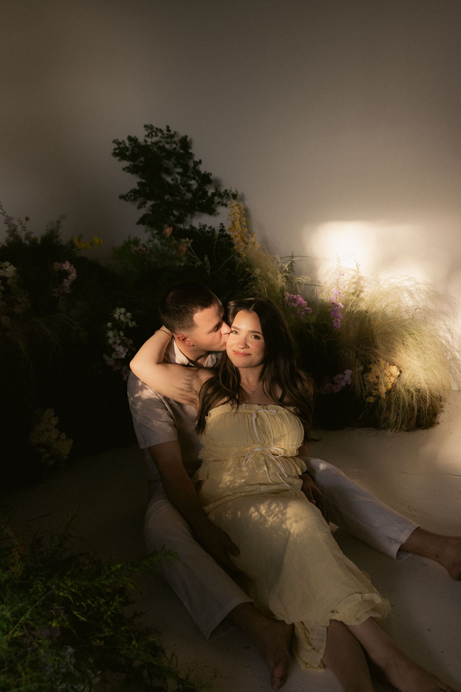 A couple sits together among lush plants, the man gently kissing the woman's cheek in warm, romantic lighting.