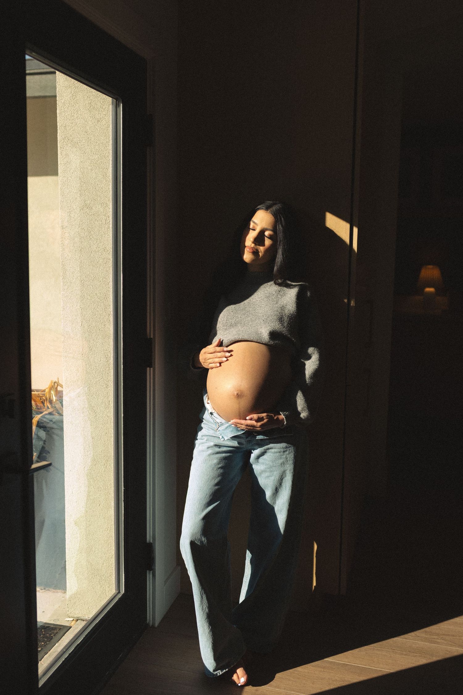 Pregnant woman in grey crop top and jeans cradling baby bump while leaning against wall in natural sunlight.