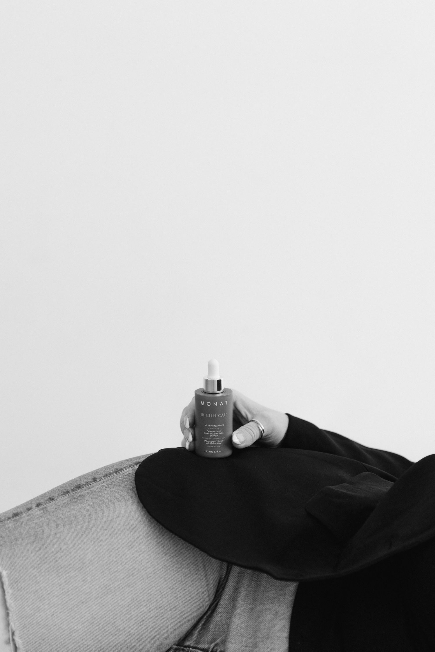 Black and white photo of a hand holding a dropper bottle serum, resting on a wide-brim hat and denim jacket.