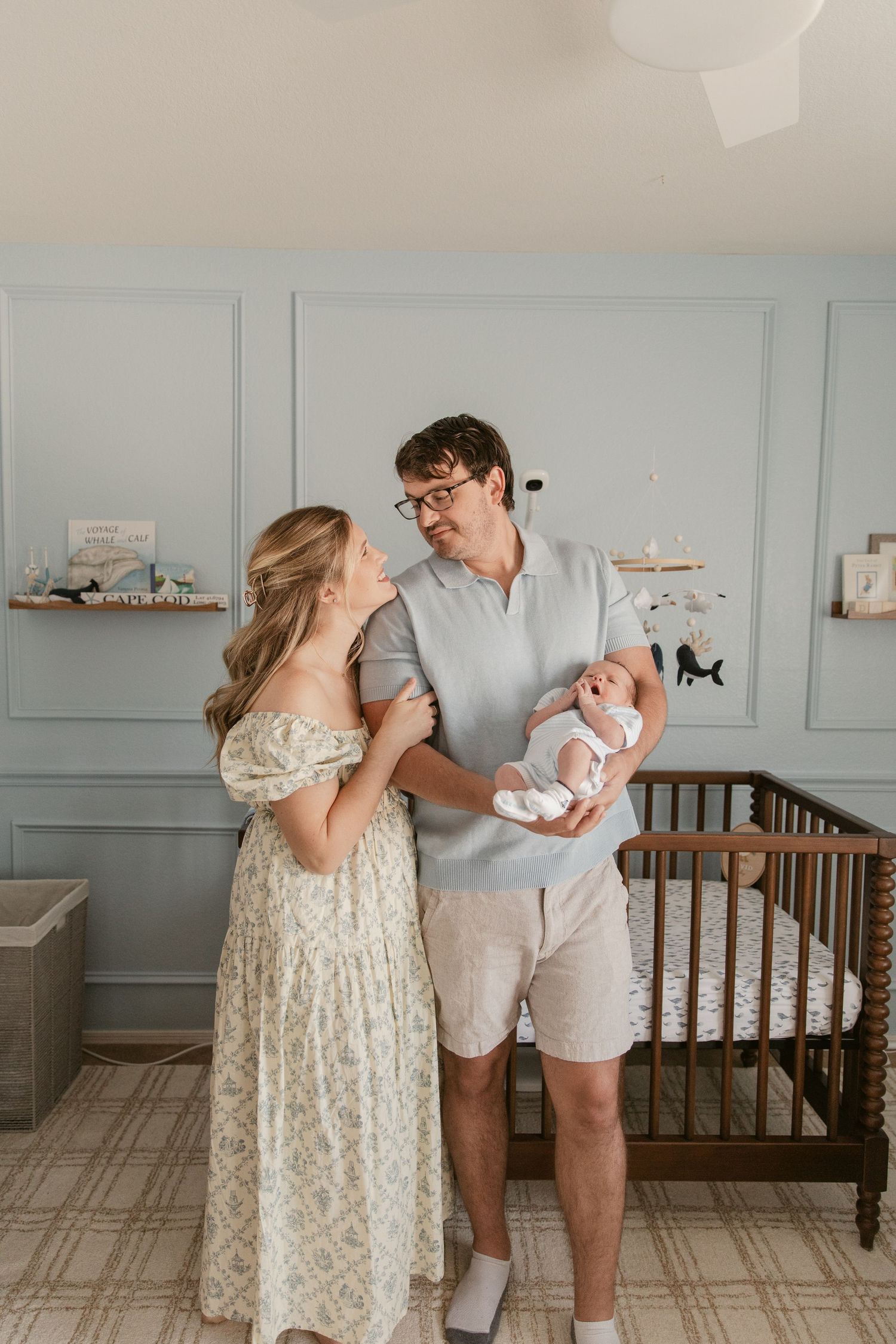 Parents share an intimate moment with their newborn baby in a nursery with gray walls and classic wooden crib.