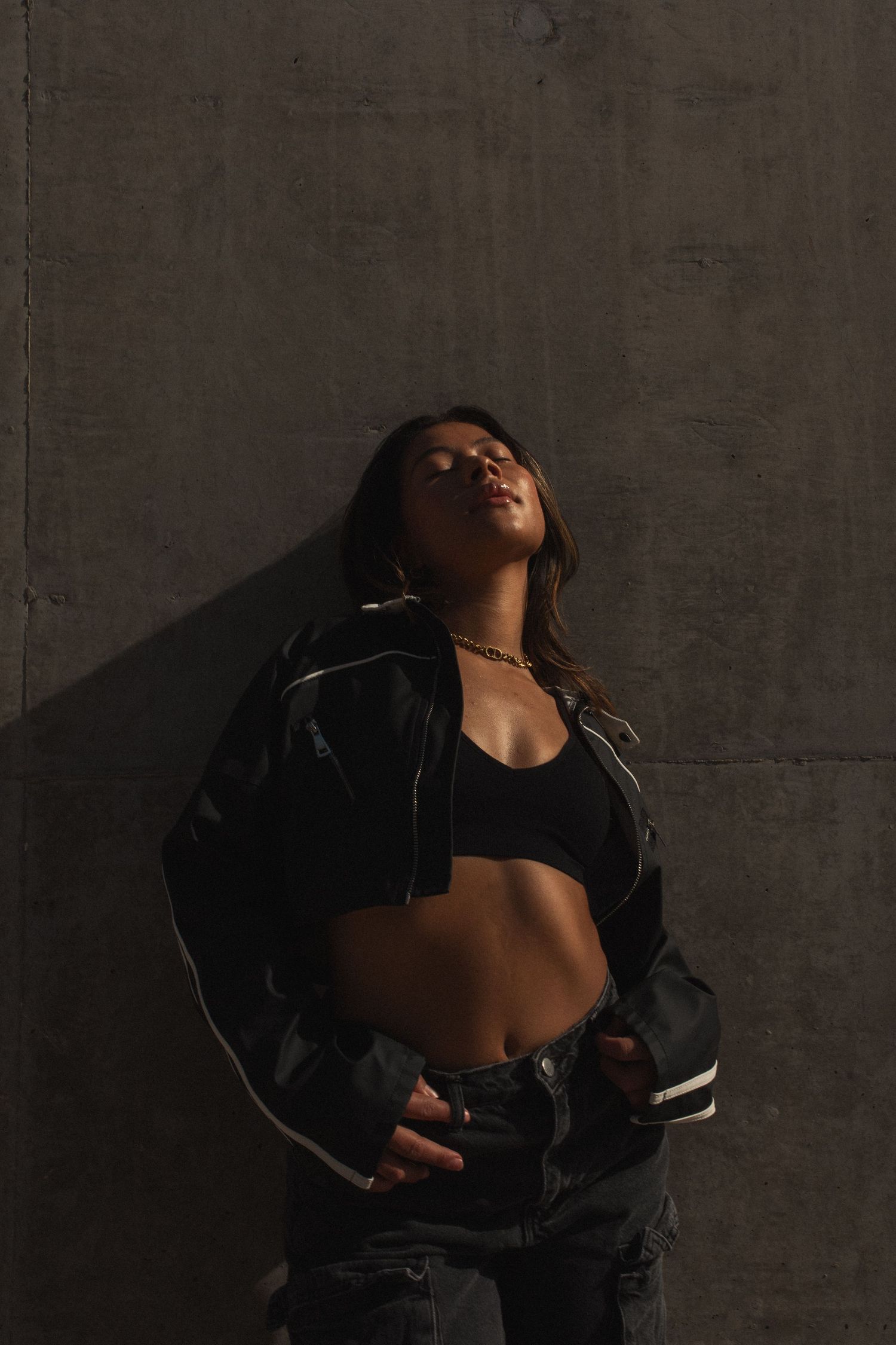 A woman in black crop top and denim jacket poses expressively against a dark concrete wall in moody lighting.
