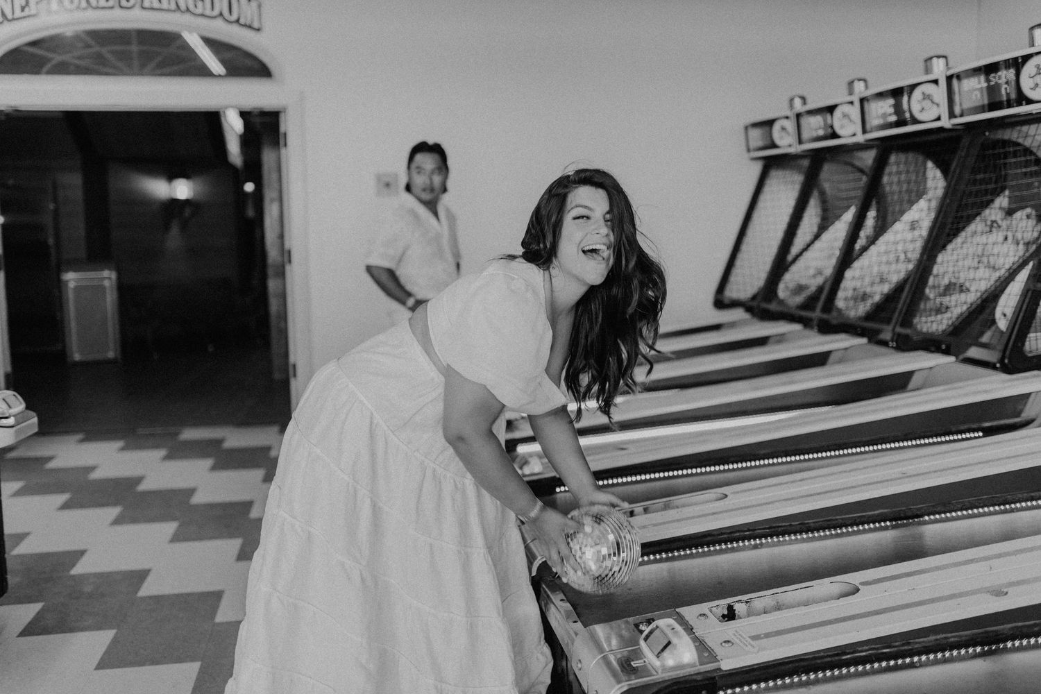 Black and white photo showing person in white dress playing vintage pinball machine in arcade.